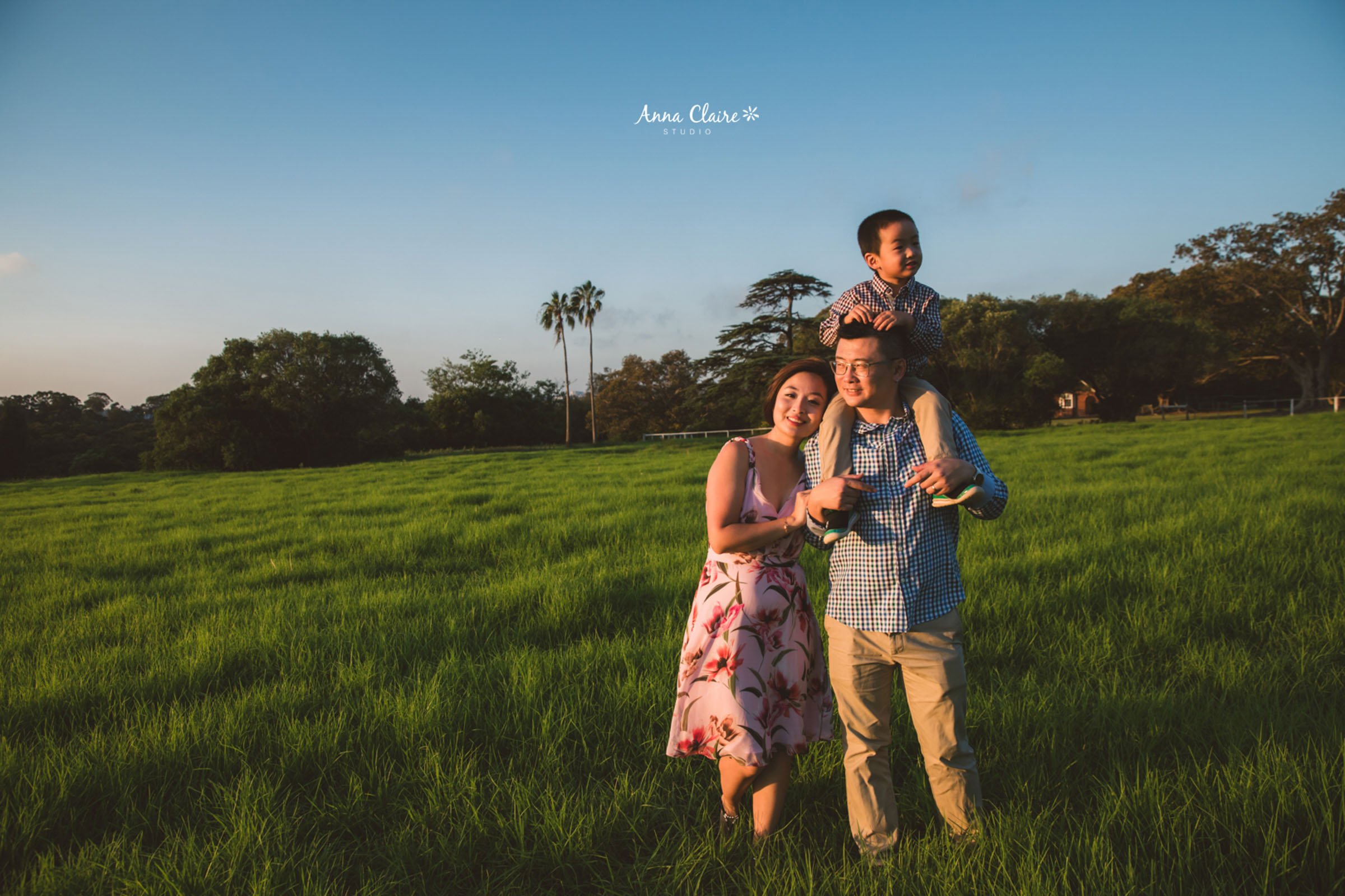 A smiling family of three, including a woman, a man, and a young boy, enjoying time together in a lush green field during sunset.