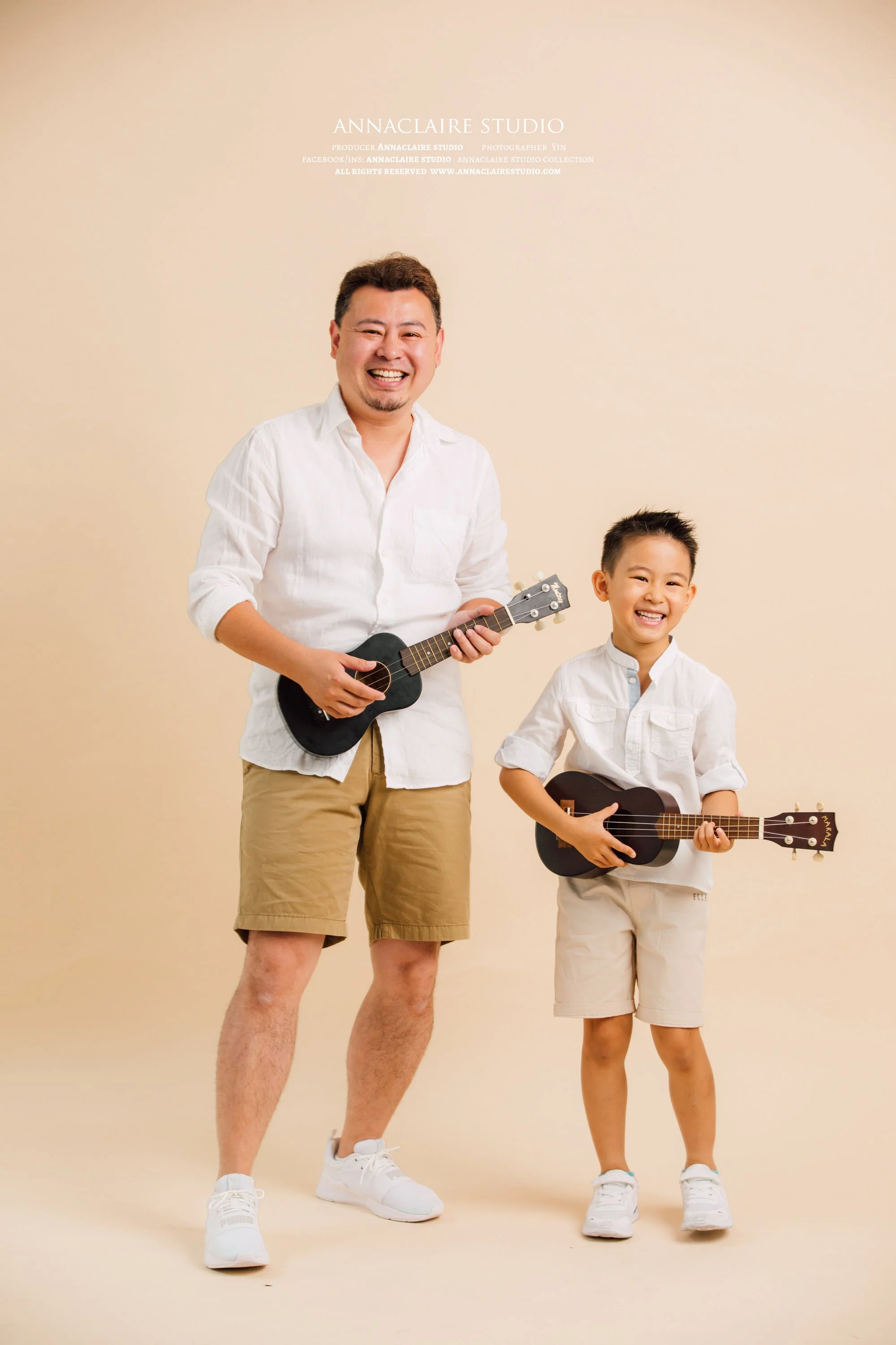 A smiling man and boy holding small black guitars against a beige background, both dressed in white shirts and beige shorts.