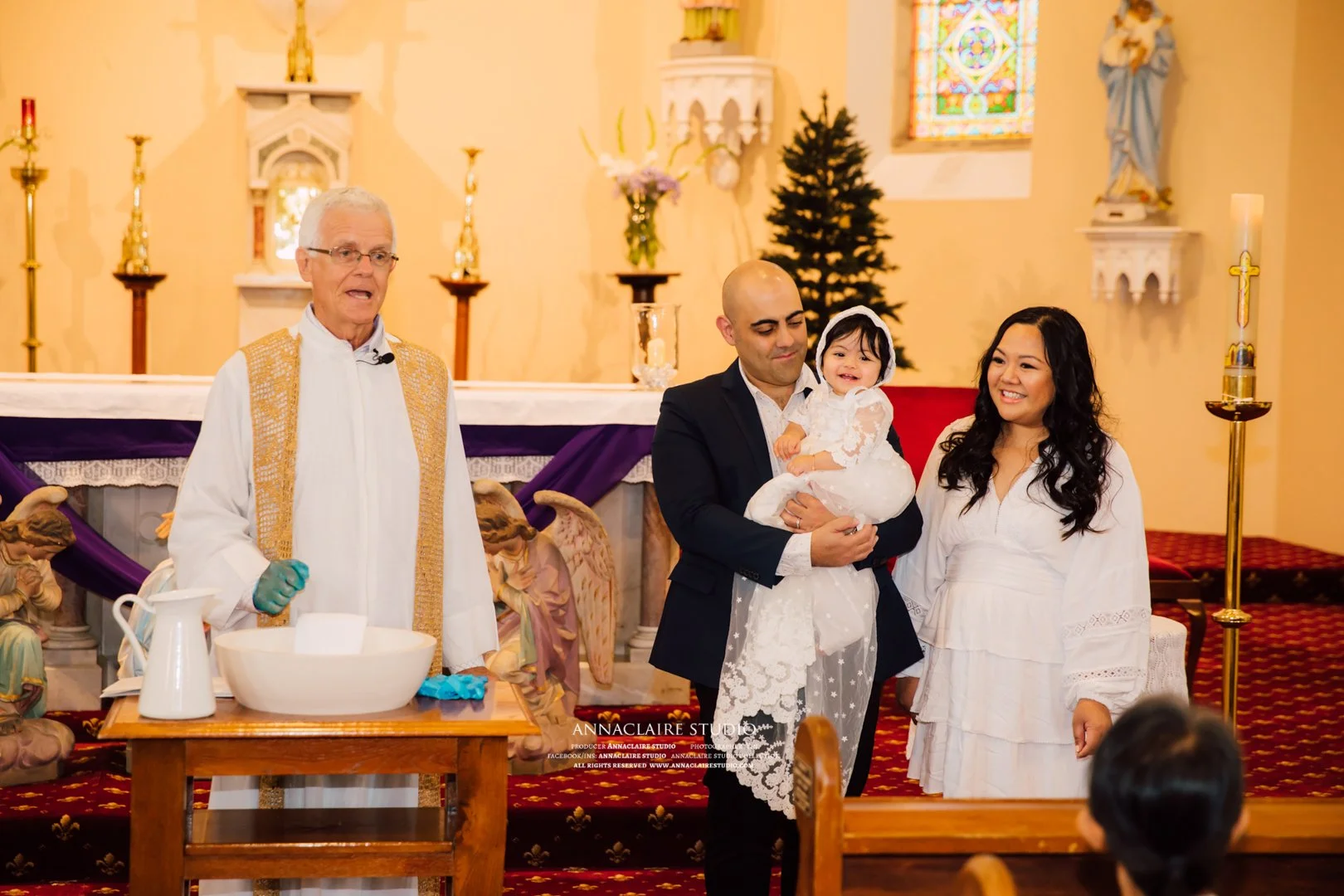 A baptism ceremony inside a church with a priest, a man holding a smiling young girl dressed in white, and a woman in a white dress standing nearby, with religious icons and decorations in the background.