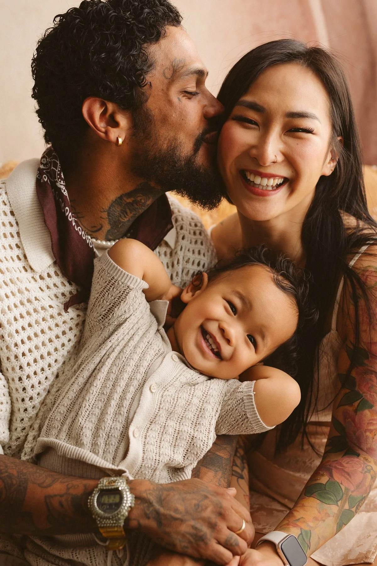 A happy family portrait of a diverse couple with their smiling toddler. The man is kissing the woman's cheek, and she is smiling broadly. The toddler is lying on the woman's lap, smiling with arms behind his head. The man has tattoos and is wearing a white knit top with a gold watch. The woman has long black hair, a nose piercing, and tattoos on her arms. The background is warm and soft.
