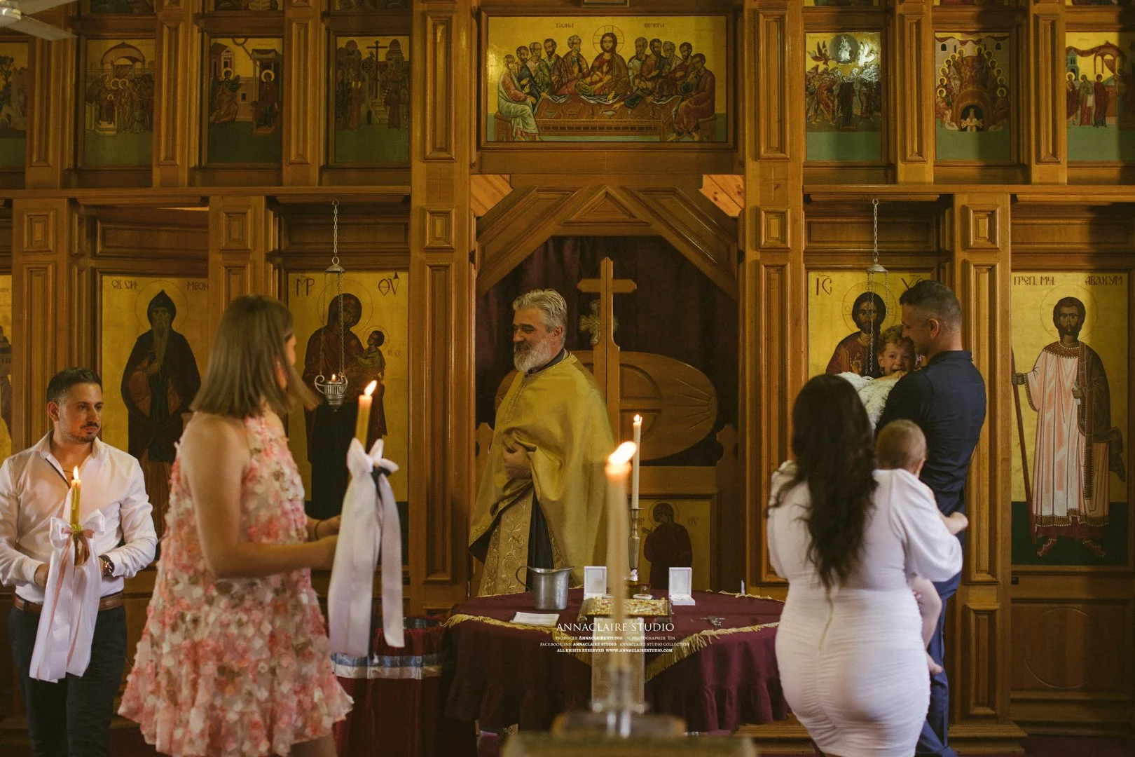 A baptism ceremony taking place inside an Orthodox church, with a priest, a man holding a child, and several other people gathered near the altar, which is decorated with lit candles and religious icons on wooden walls.