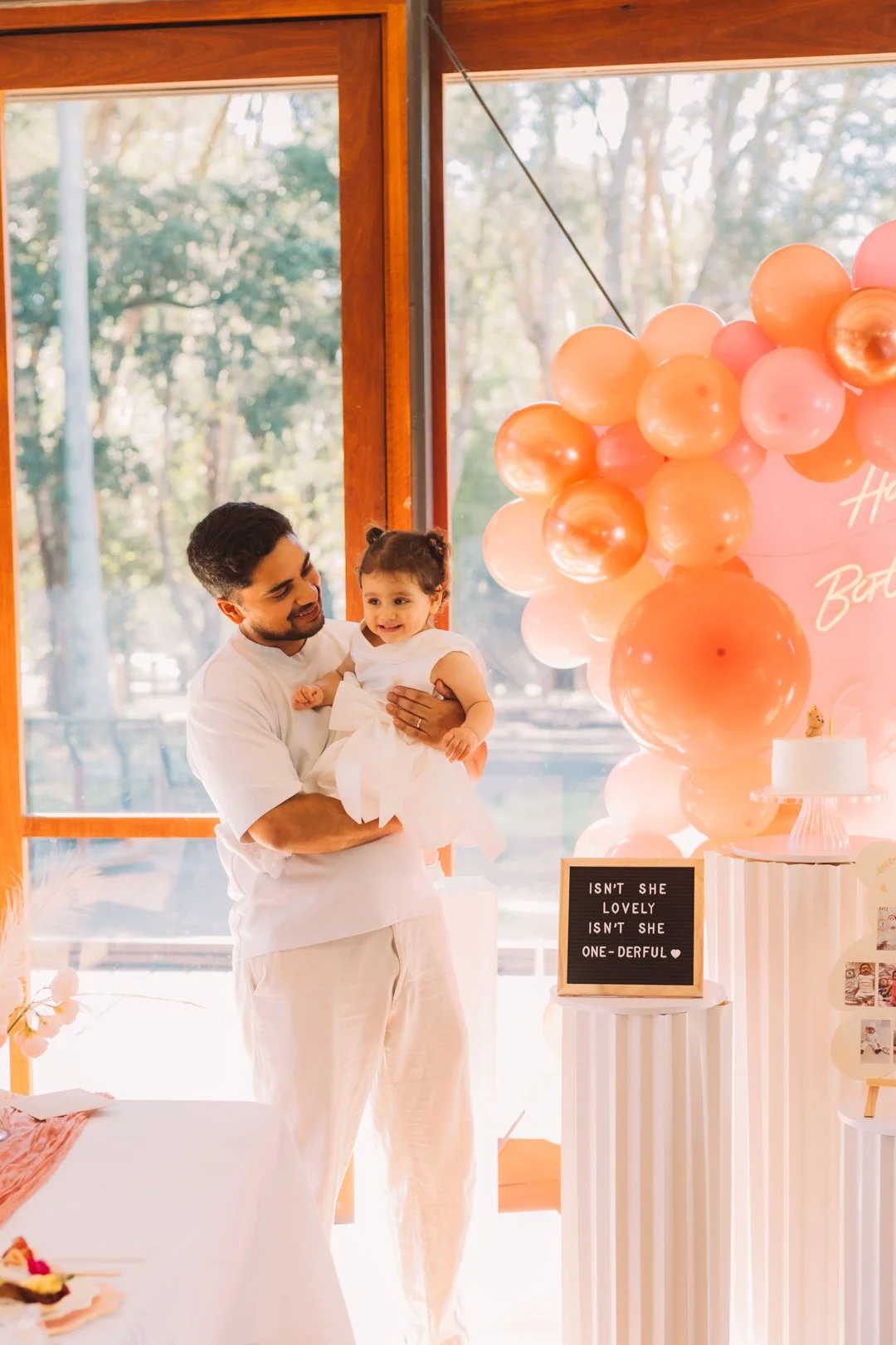 A man holding a smiling young girl dressed in white at a pink-themed birthday party with balloons, a cake, and a sign that reads 'Isn't she lovely isn't she one-derful'.