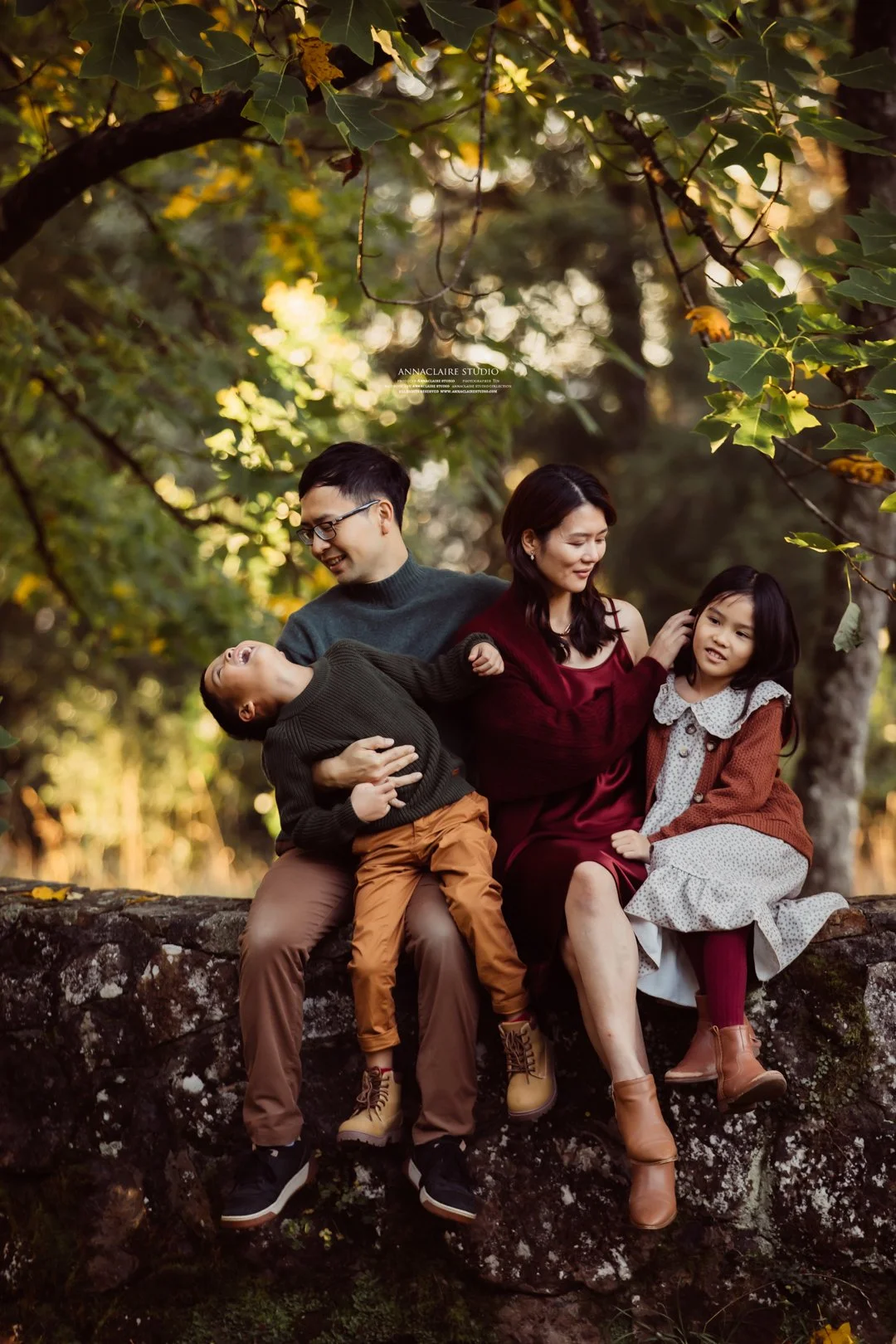 A family of four sitting on a moss-covered rock in a wooded area with green leaves and sunlight filtering through the trees. The father, mother, and two children are smiling and interacting with each other.