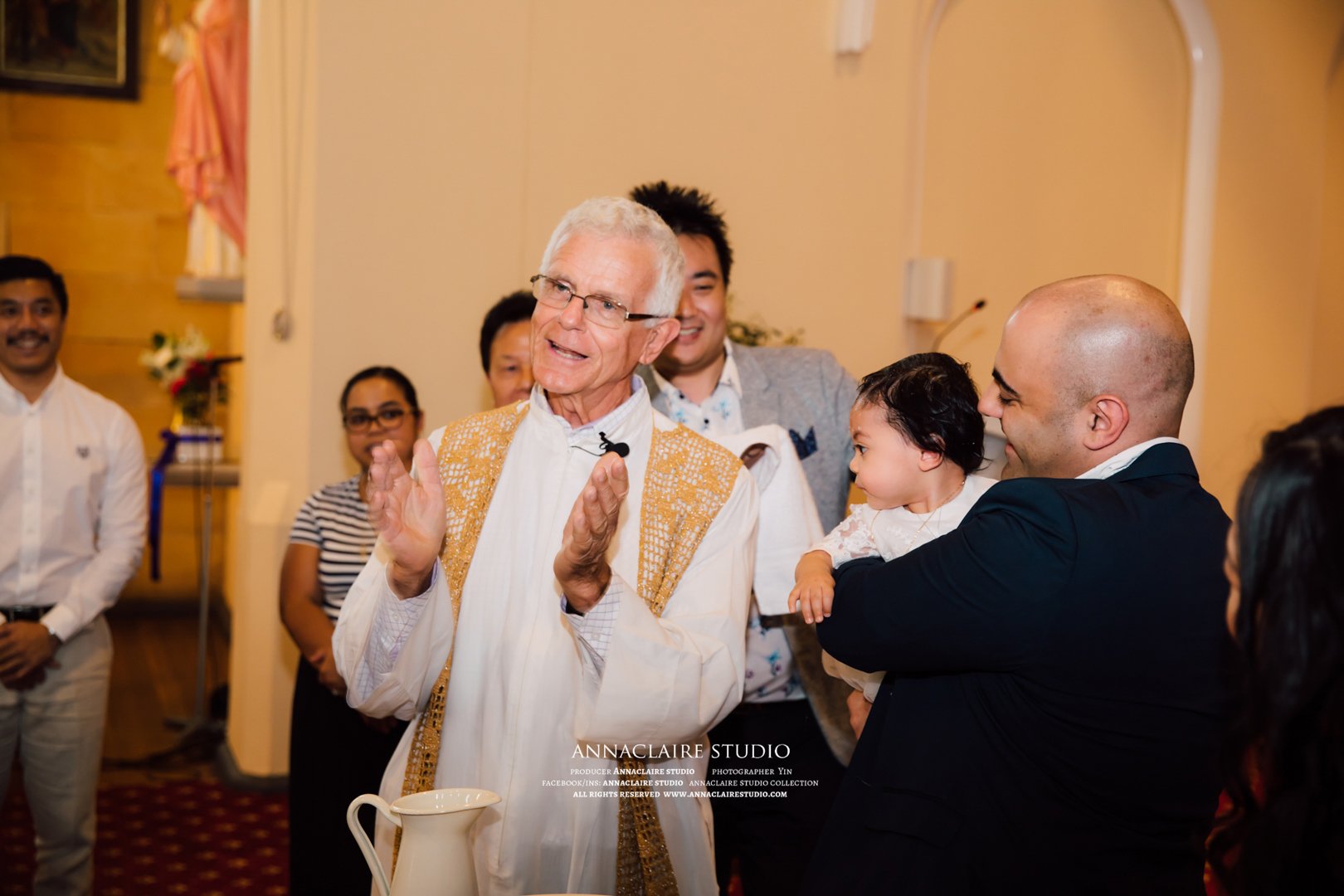 A group of people gathered around a priest during a celebration. The priest is clapping and smiling, with a pitcher on a table in the foreground. A man holds a young girl, both smiling, and other attendees are standing and smiling in the background.