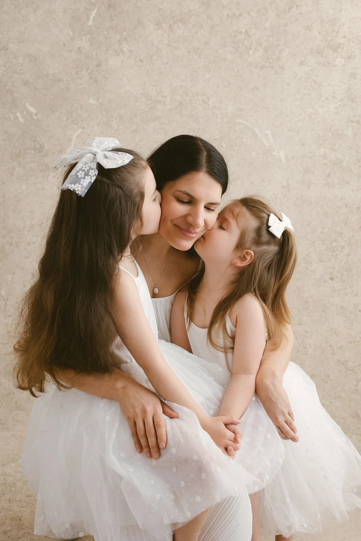 A woman with two young girls, all dressed in white, sharing a loving hug and kisses.