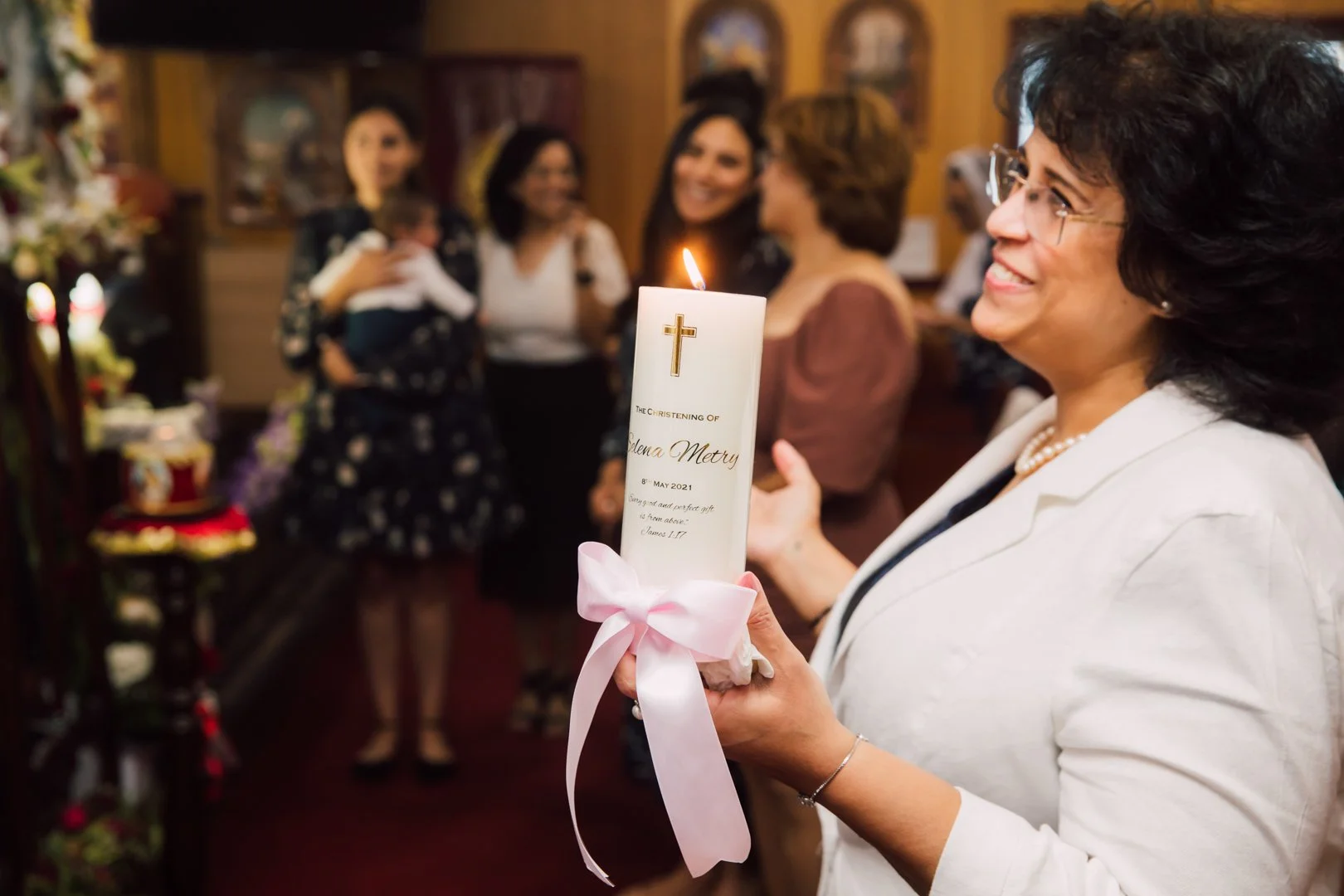A woman is holding a lit candle with a cross symbol and text about a christening, with a group of women, one holding a baby, gathered in a warmly decorated room celebrating a christening.