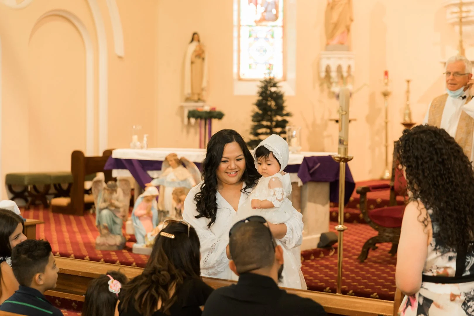 A woman holding a small girl dressed in white inside a church during a religious ceremony with children and adults present.