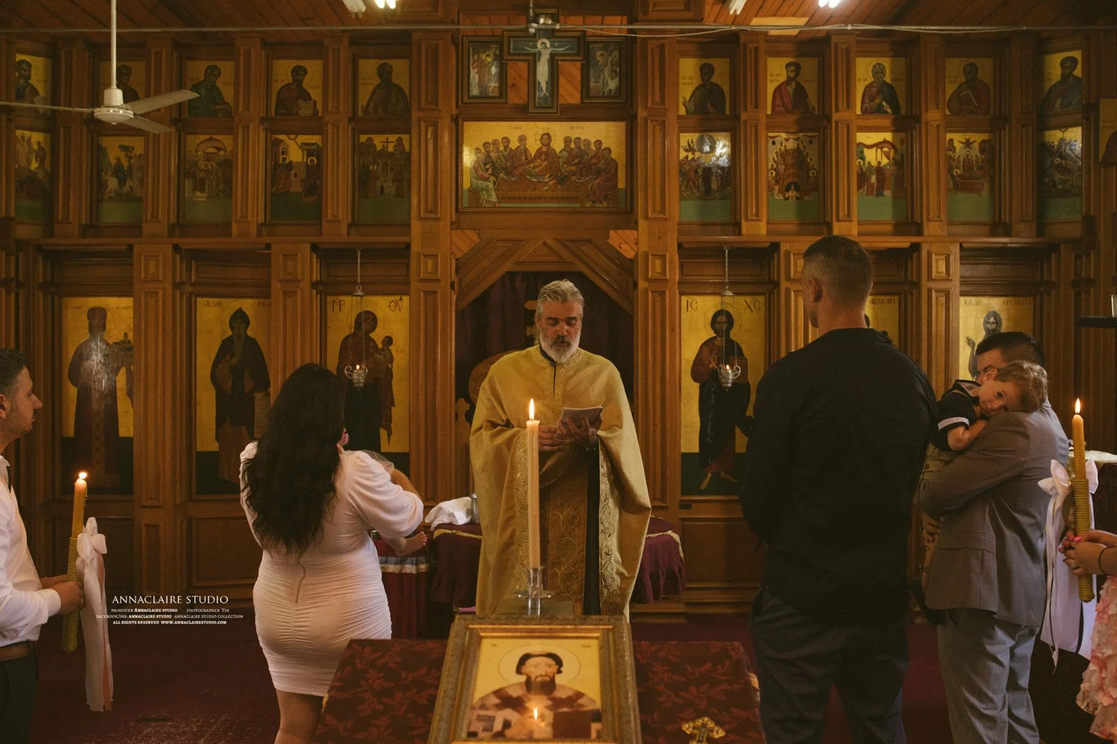 A religious ceremony inside a church with people gathered around a priest in yellow robes. The church has wooden walls with numerous icons and religious artwork. Candles are lit on the table in front of the priest, and a framed icon is visible on the table.