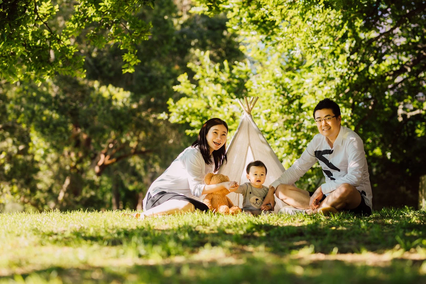 A family enjoying a picnic outdoors under trees, sitting on the grass in front of a small teepee tent, with a teddy bear and a toddler between them.