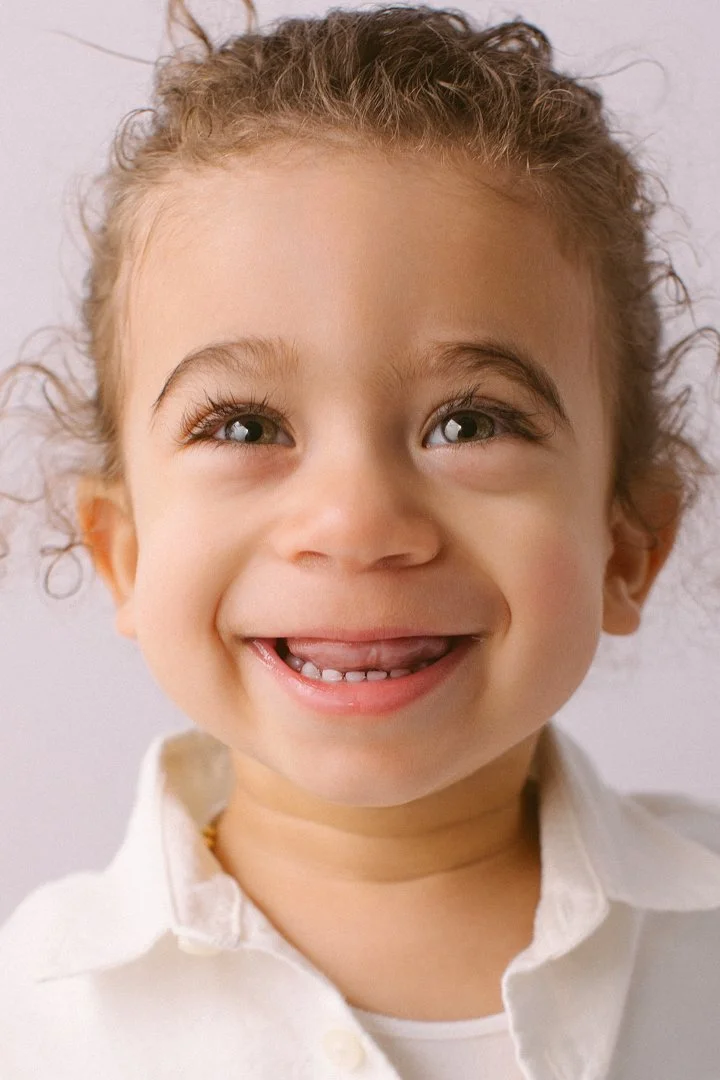Close-up of a young girl smiling and sticking out her tongue, with curly hair and bright eyes, wearing a white shirt.