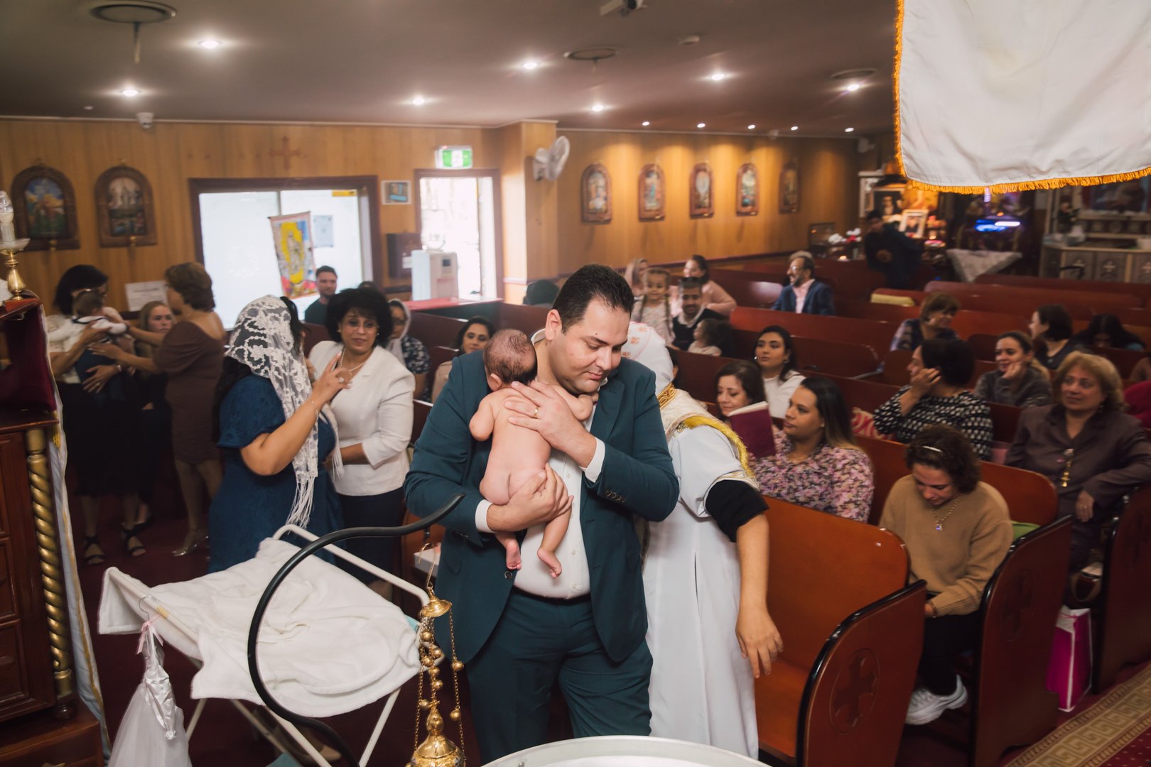 A man in a suit holding a baby during a baptism ceremony in a church with many women and children in attendance.
