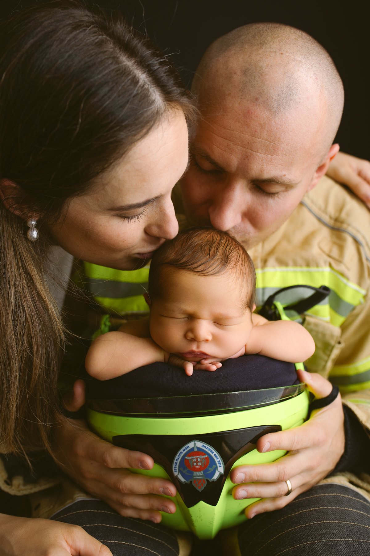 A firefighter is holding a sleeping baby in a helmet while a woman and firefighter kiss his head.