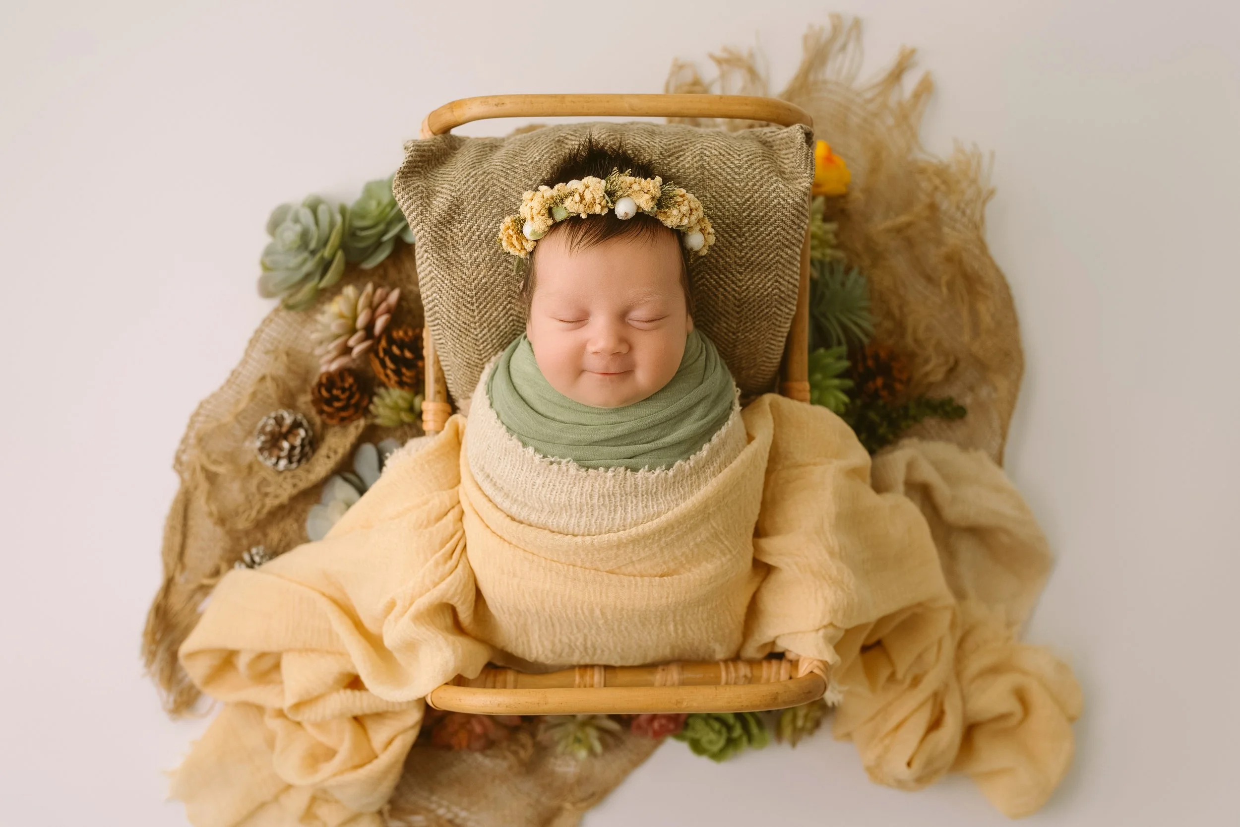 A smiling baby wearing a floral headband, wrapped in layered blankets, lying in a wicker basket surrounded by greenery and pinecones.