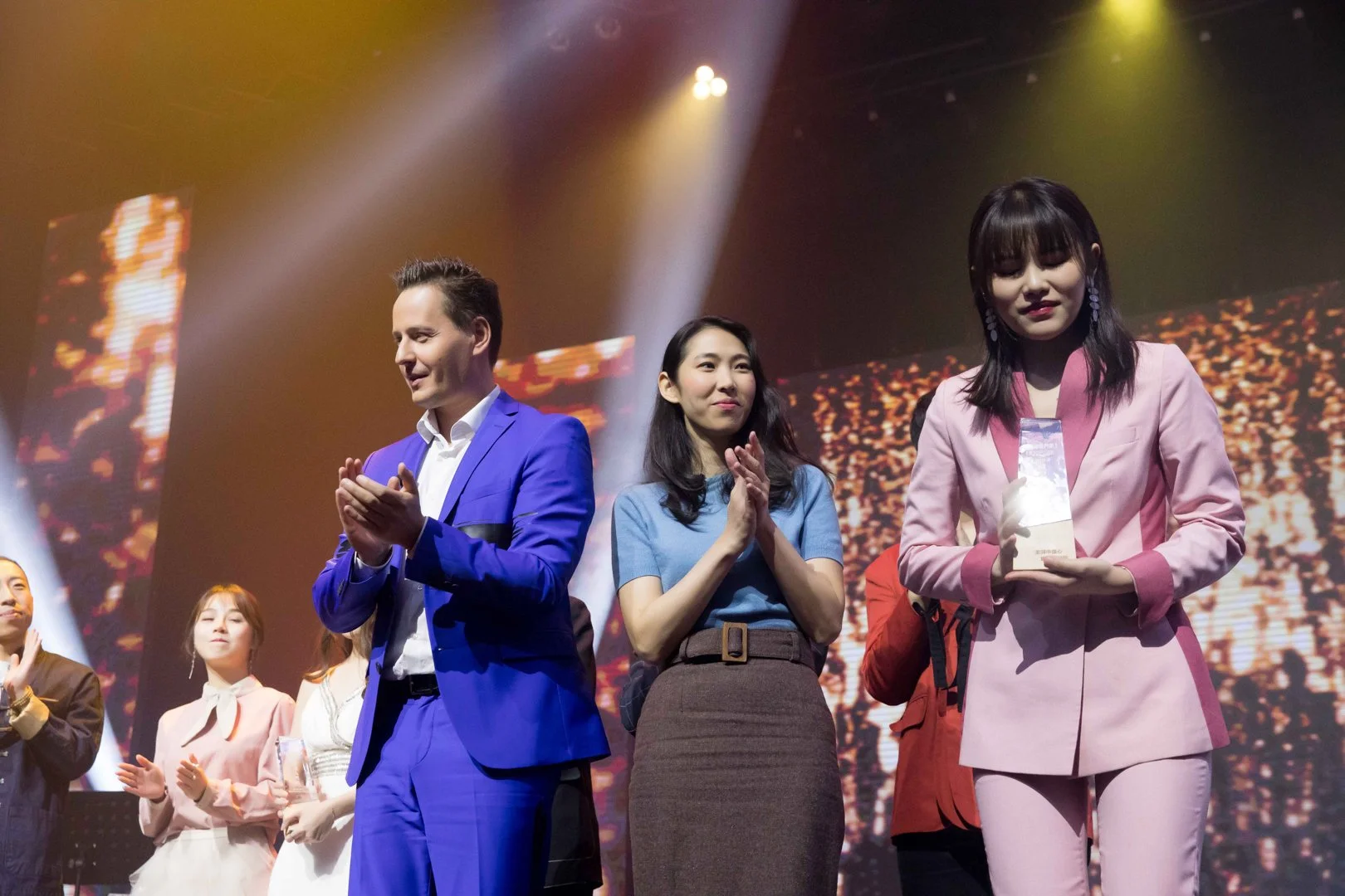 Three individuals on stage during an event, with a large screen displaying abstract visuals behind them. The man on the left is wearing a bright blue suit, the woman in the middle is dressed in a blue top and brown skirt, and the woman on the right i