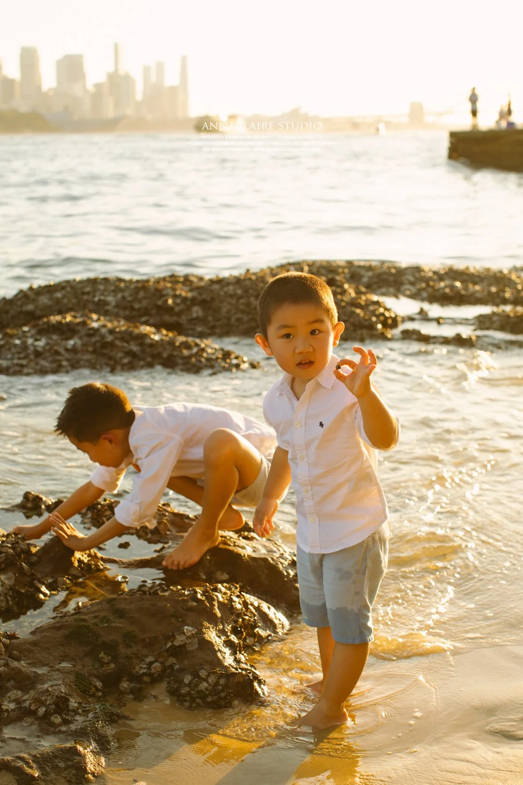 Two young boys in white shirts and shorts playing by the beach during sunset, one is touching the water and the other is climbing on rocks with a Sydney city skyline in the background.