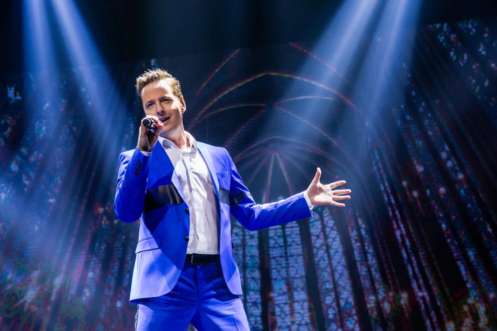 A man wearing a blue suit and white shirt is singing into a microphone on stage with purple and blue lighting, in front of a backdrop resembling a stained glass window.