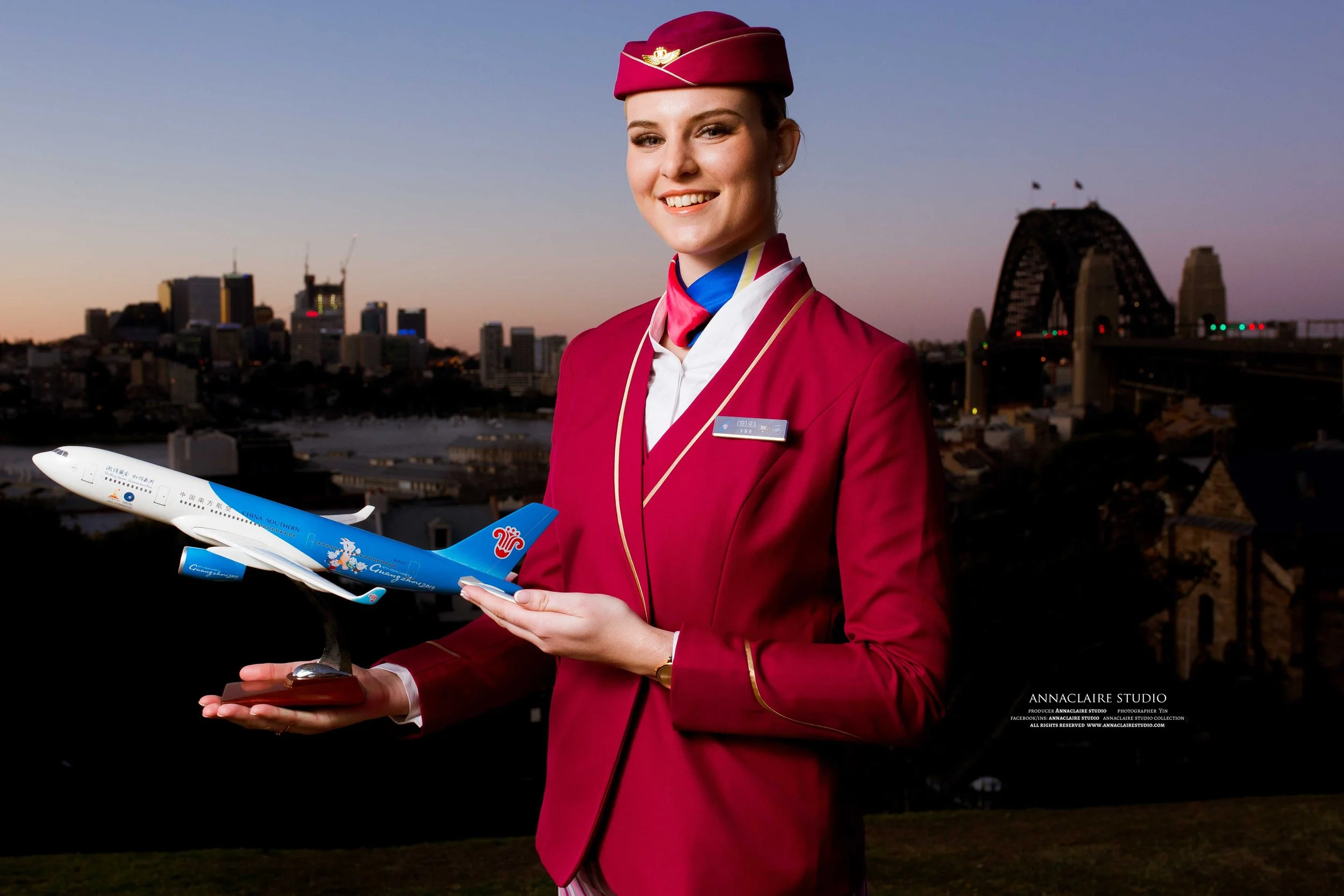 A smiling flight china southern airlines female flight attendant in a red uniform and matching hat holding a model airplane with a cityscape and bridge in the background during sunset.