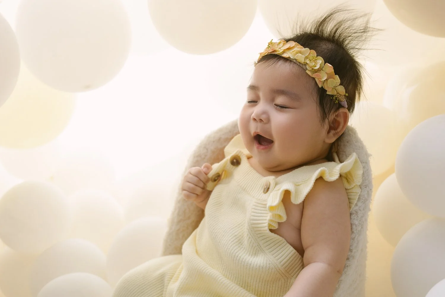 A smiling baby girl with a yellow headband and cream-colored outfit surrounded by white balloons.
