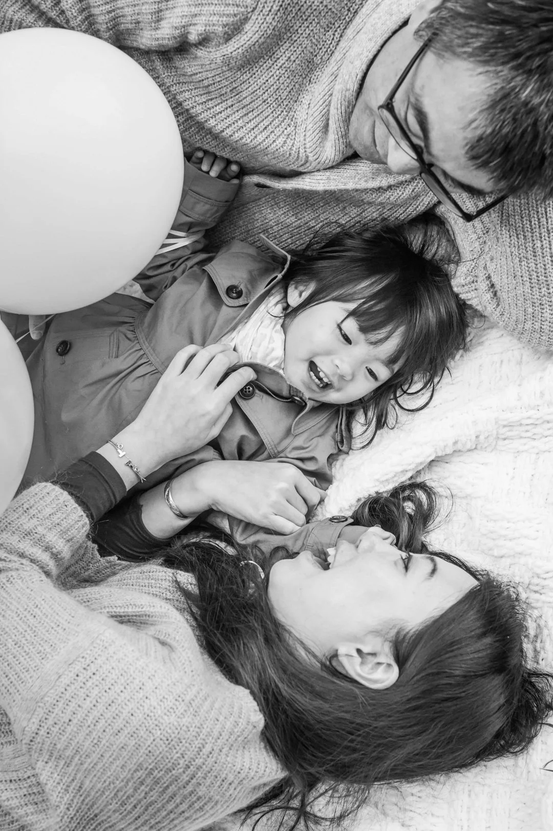 A woman and a young girl lying on a bed with a woman, a girl, and an older man, smiling and interacting while holding balloons nearby. The image is in black and white.