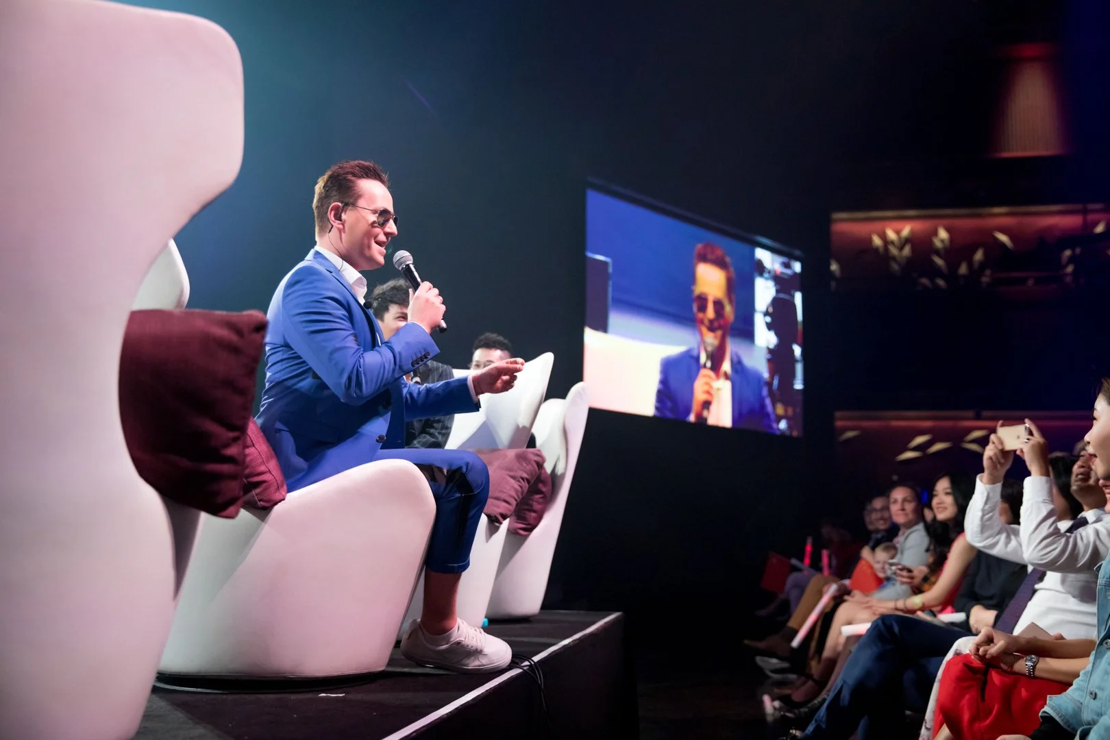 A man in a blue suit and sunglasses is sitting on a white chair on stage, holding a microphone and smiling. He is facing an audience and a large screen displays his reflection in the background.