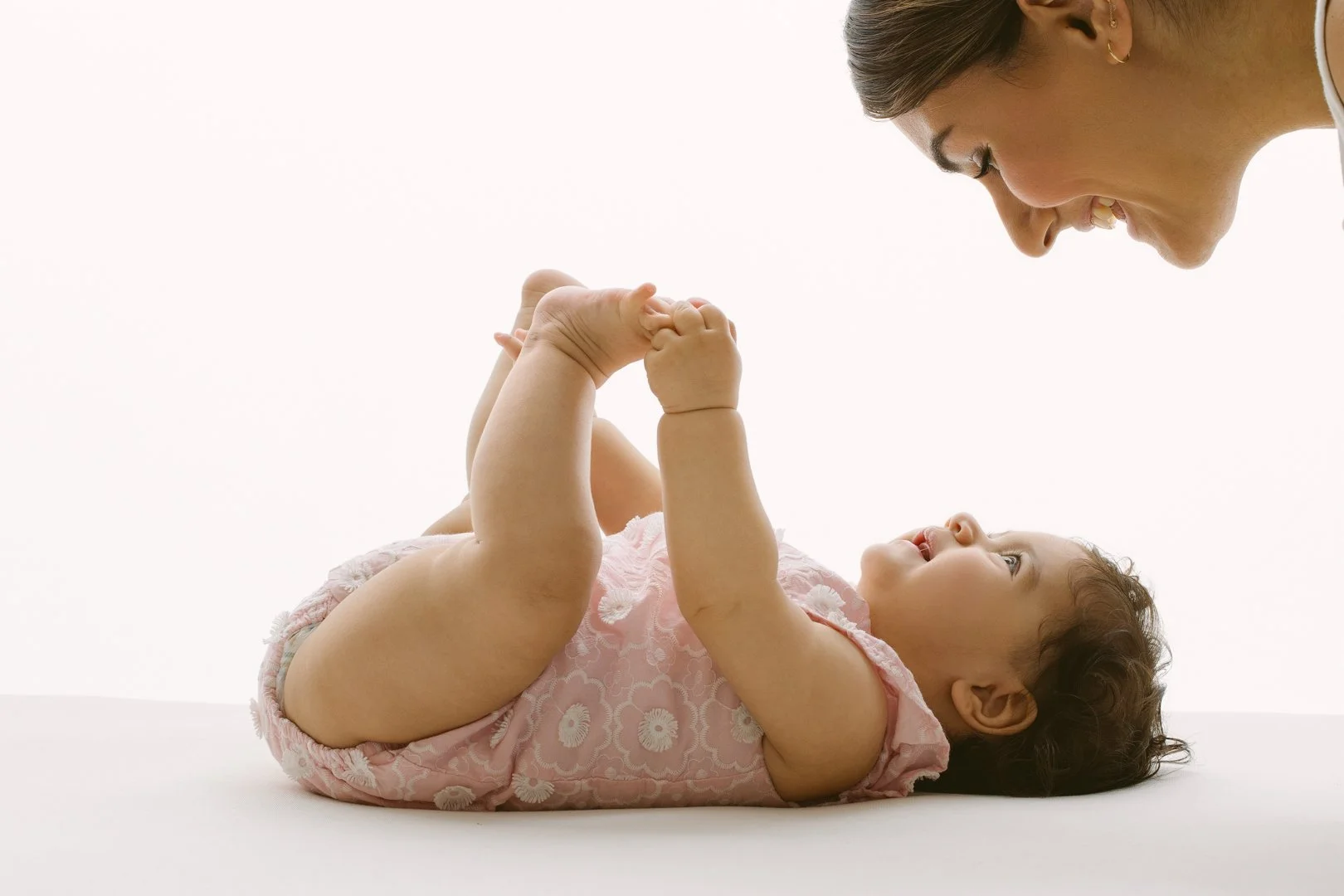 A baby lying on back, holding its feet, and smiling up at a woman who is leaning over and making eye contact with the baby.