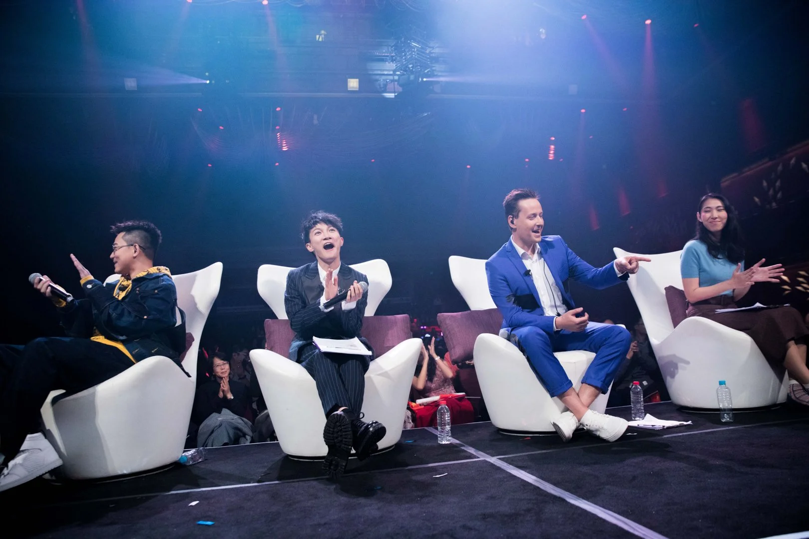 Four people sitting on white chairs on stage during a live event, engaging in conversation and smiling. The man second from the right in a blue suit is pointing, and the other three are clapping or holding microphones, with an audience visible in the