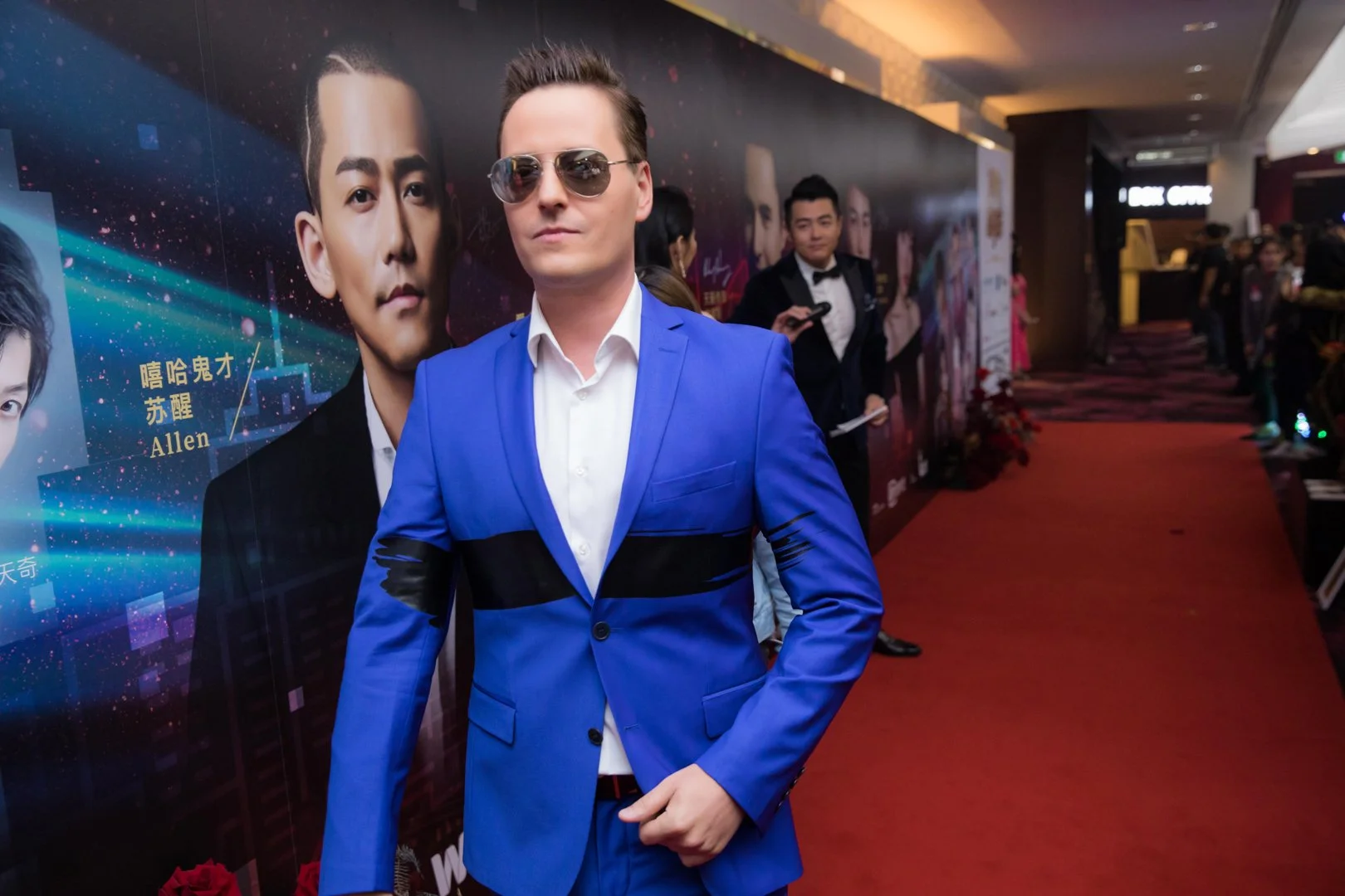 A man in a bright blue suit, white shirt, and sunglasses standing on a red carpet at an event, with a wall of promotional posters behind him.