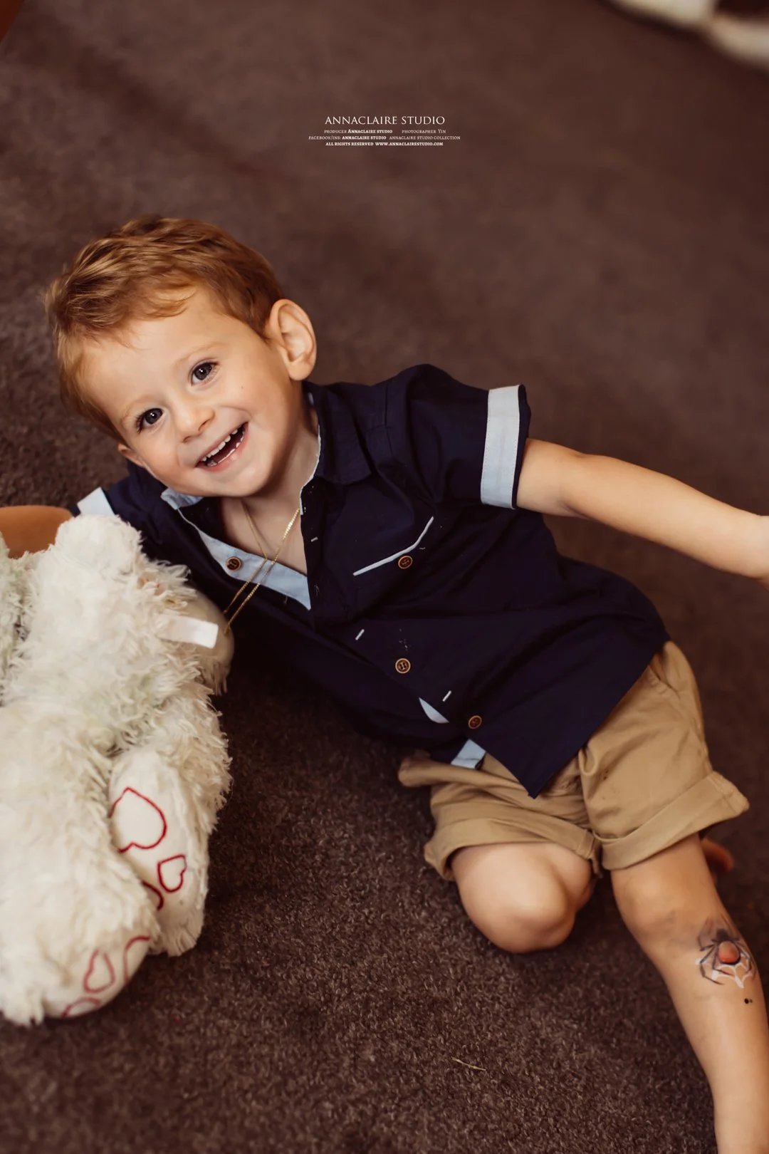 A young boy with red hair smiling and lying on a brown carpet next to a plush teddy bear. The boy is wearing a navy blue shirt and beige shorts, with a tattoo on his lower leg.