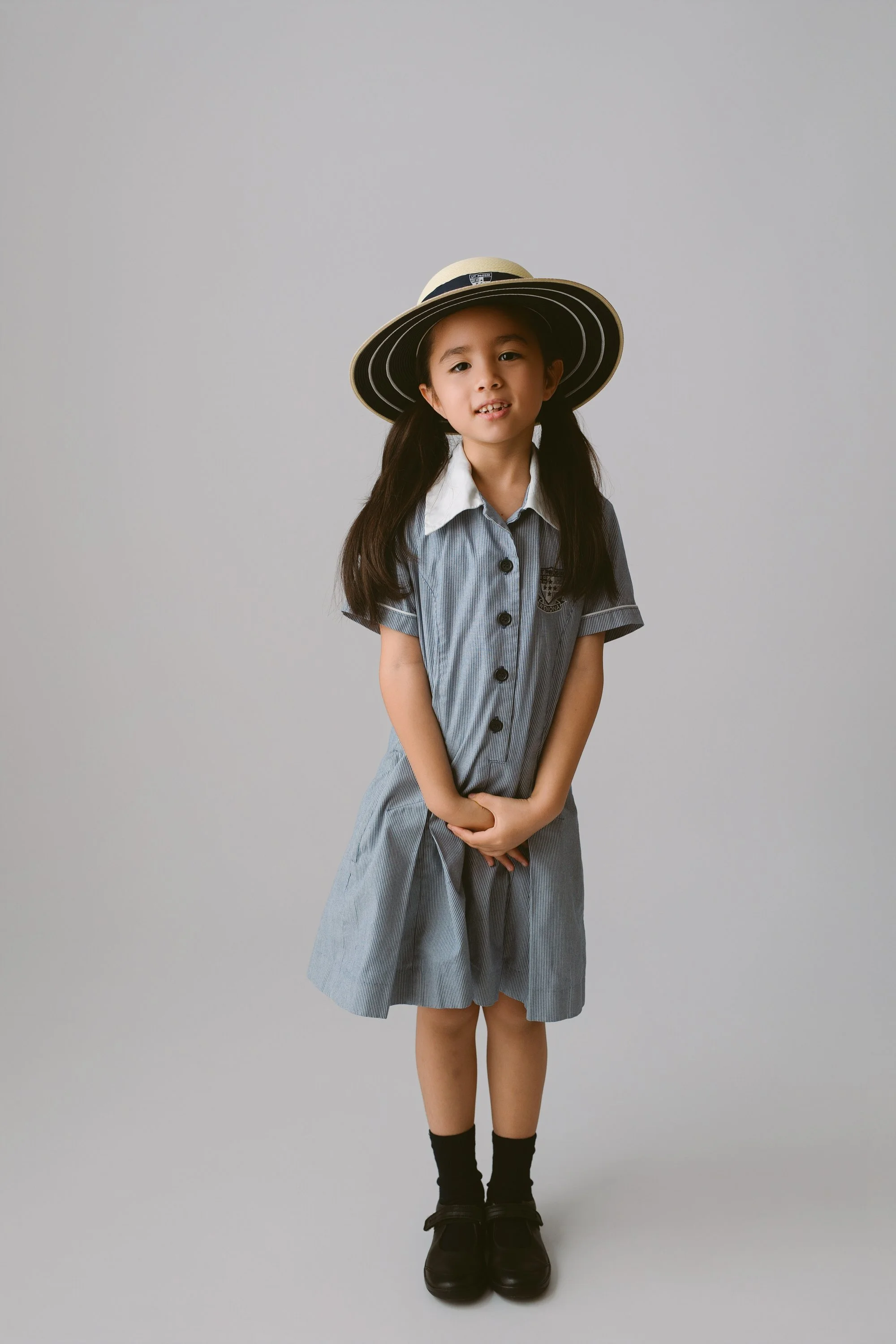 Young girl dressed in a school uniform with a striped dress, white collar, and black shoes, wearing a wide-brimmed hat, standing against a plain background.
