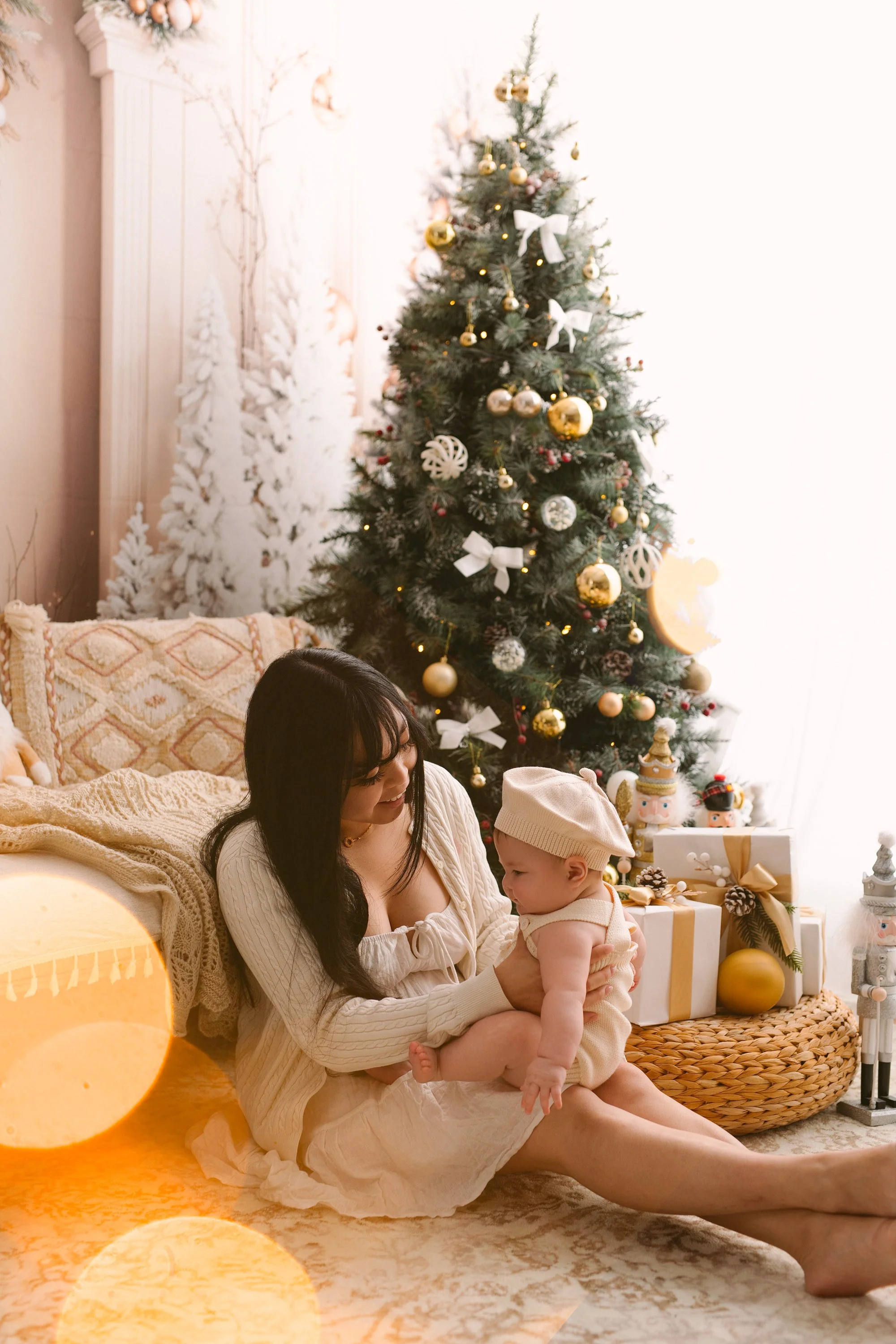 A woman sitting on the floor with a baby, near a decorated Christmas tree and wrapped presents, during Christmas.