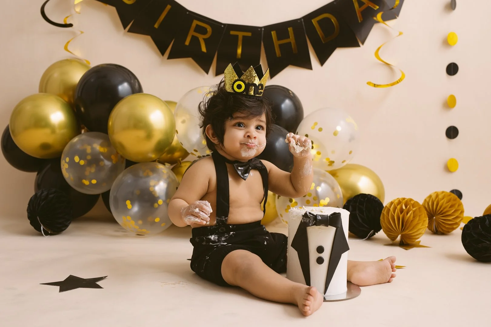 A young child celebrating a first birthday, sitting on the floor with a cake. The child is wearing a black bow tie, suspenders, and a gold crown with the number "one" on it. The background is decorated with black, gold, and transparent balloons, and black and gold paper decorations.