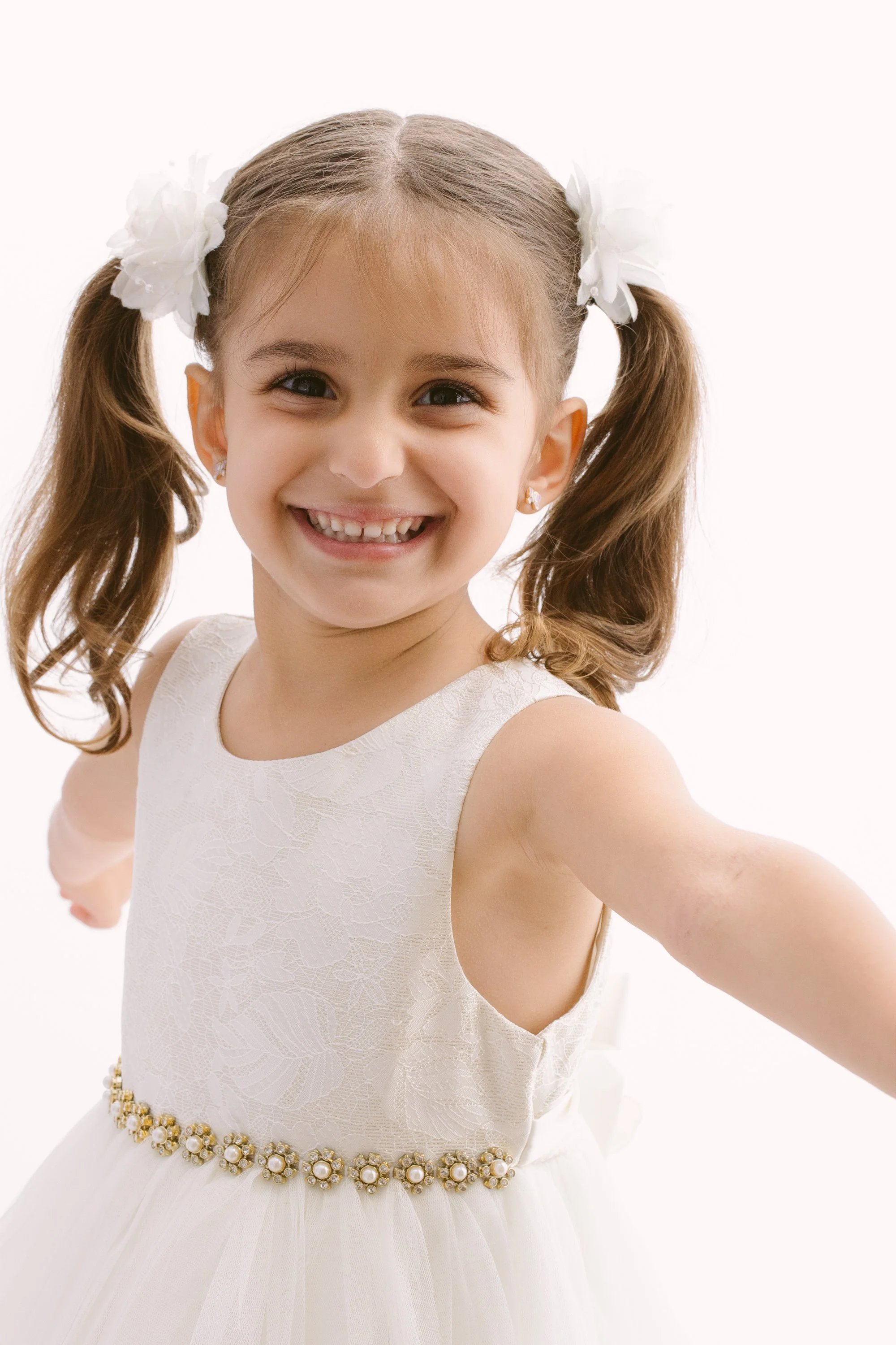 A young girl smiling, wearing a white dress with floral embroidery and pearl embellishments at the waist, and white flower hair accessories.
