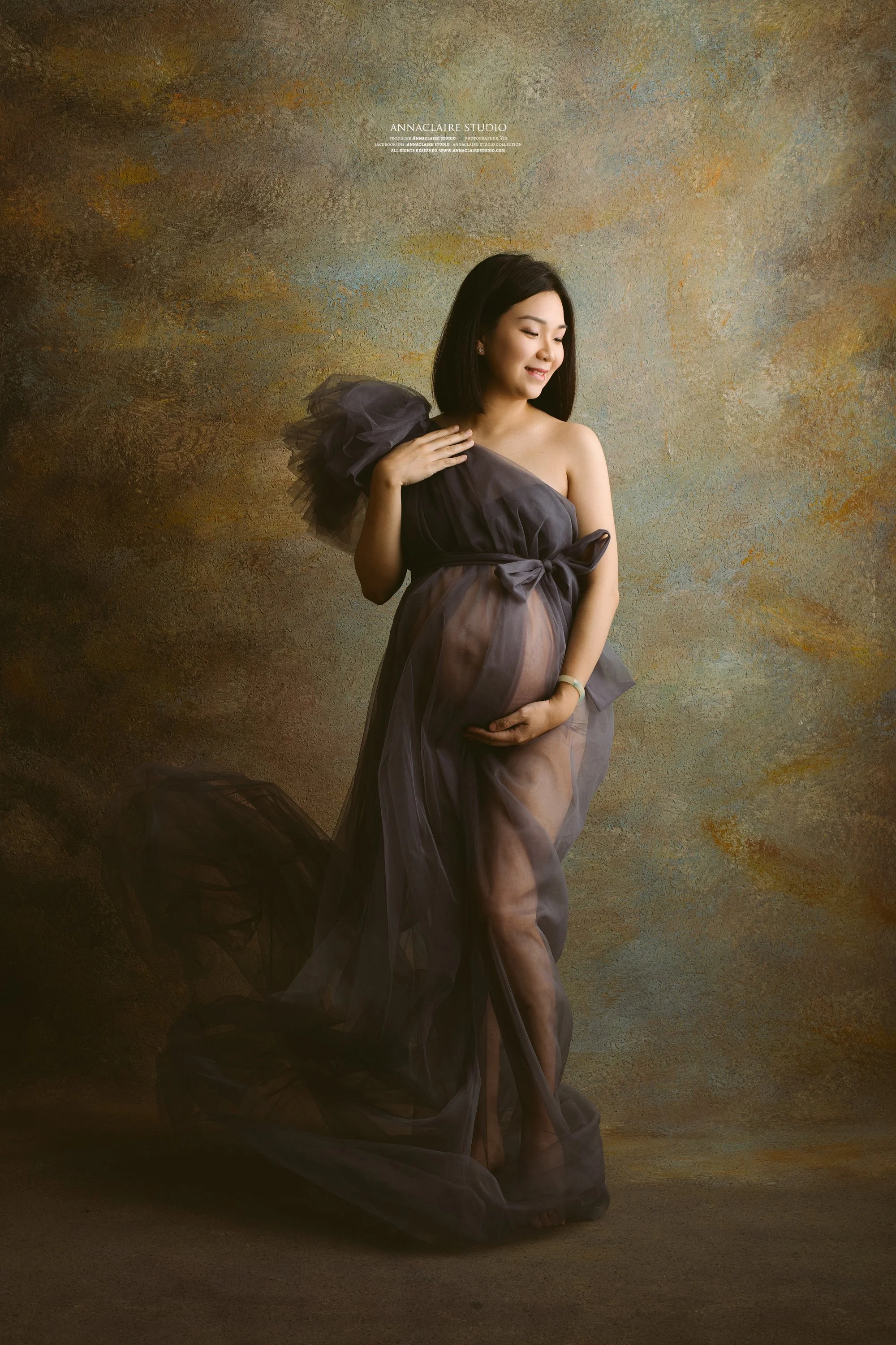 Pregnant woman in a sheer black gown posing against a textured brown backdrop.