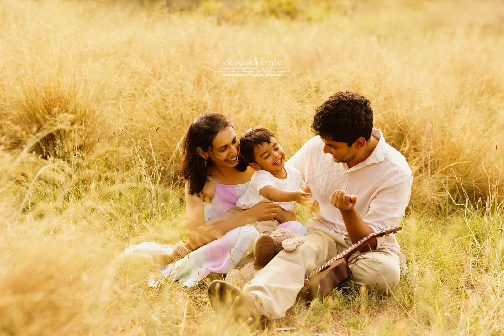 A family of three sitting in a field of tall yellow grass, smiling and playing together during sunset at Centennial Park .