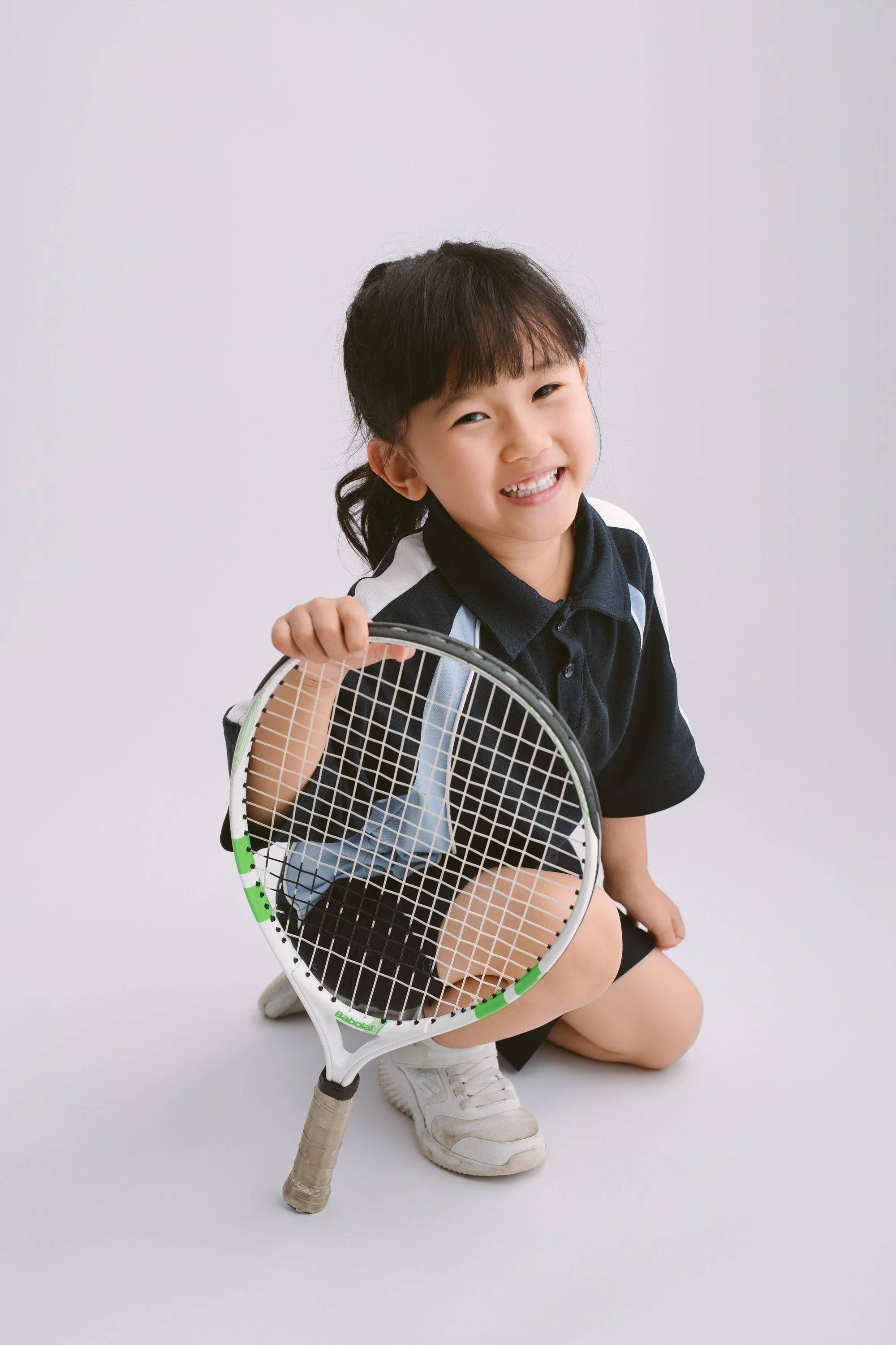 A young girl crouching on the floor holding a tennis racquet, smiling at the camera, wearing a sports shirt and sneakers.
