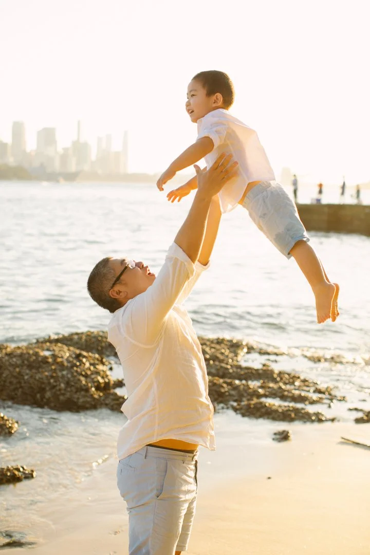 A man lifting a young boy in the air at the beach during sunset with city Sydney skyline in the background.