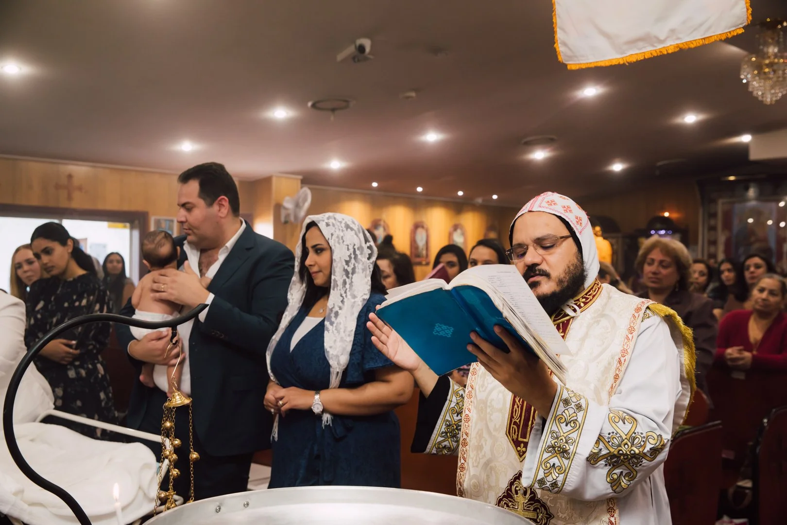 An Orthodox Christian baptism ceremony inside a church, with a priest reading from a religious book, a man holding a baby, and a congregation of worshippers in the background.