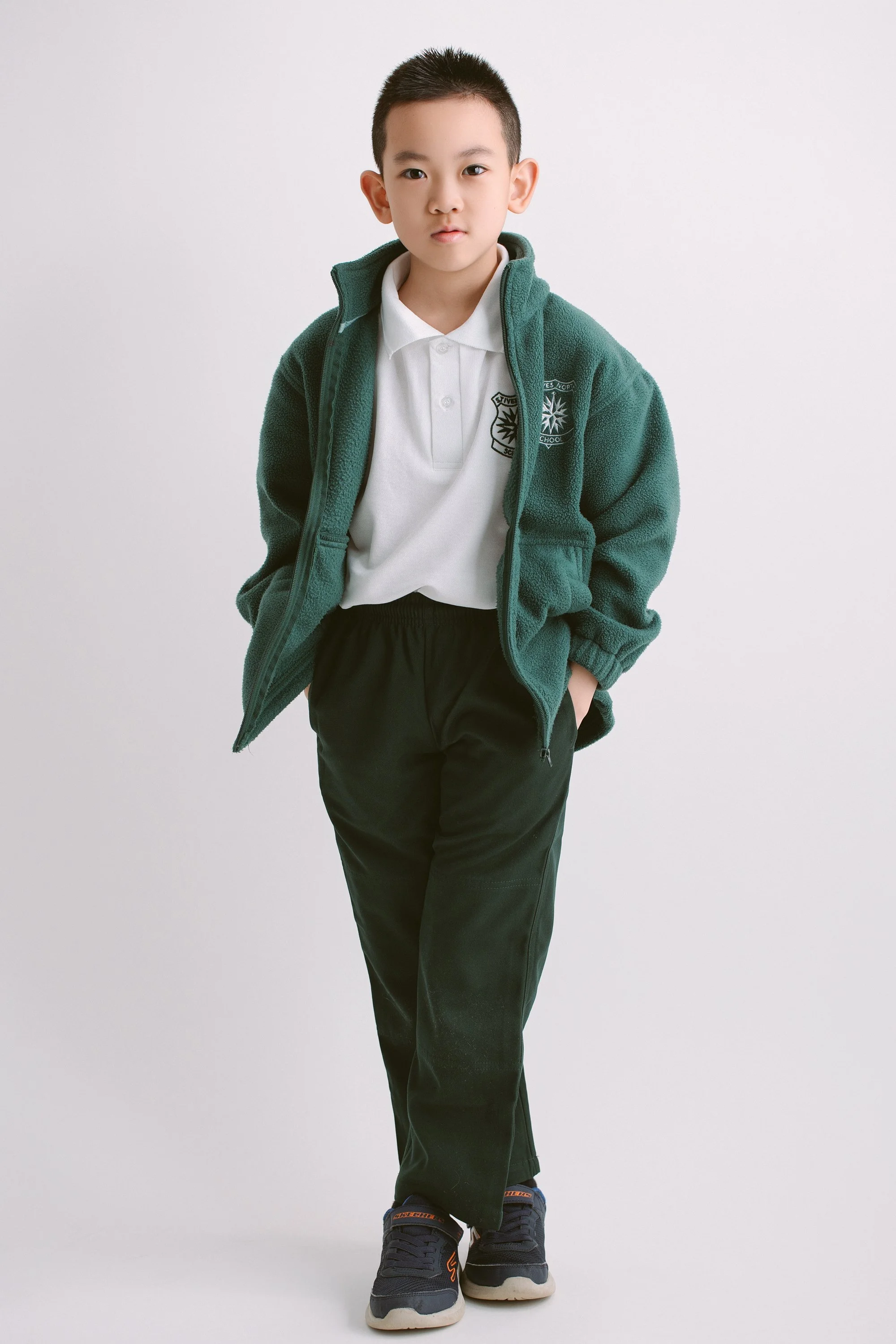 A young boy wearing a white polo shirt, green fleece jacket, black sweatpants, and sneakers, standing against a plain white background.