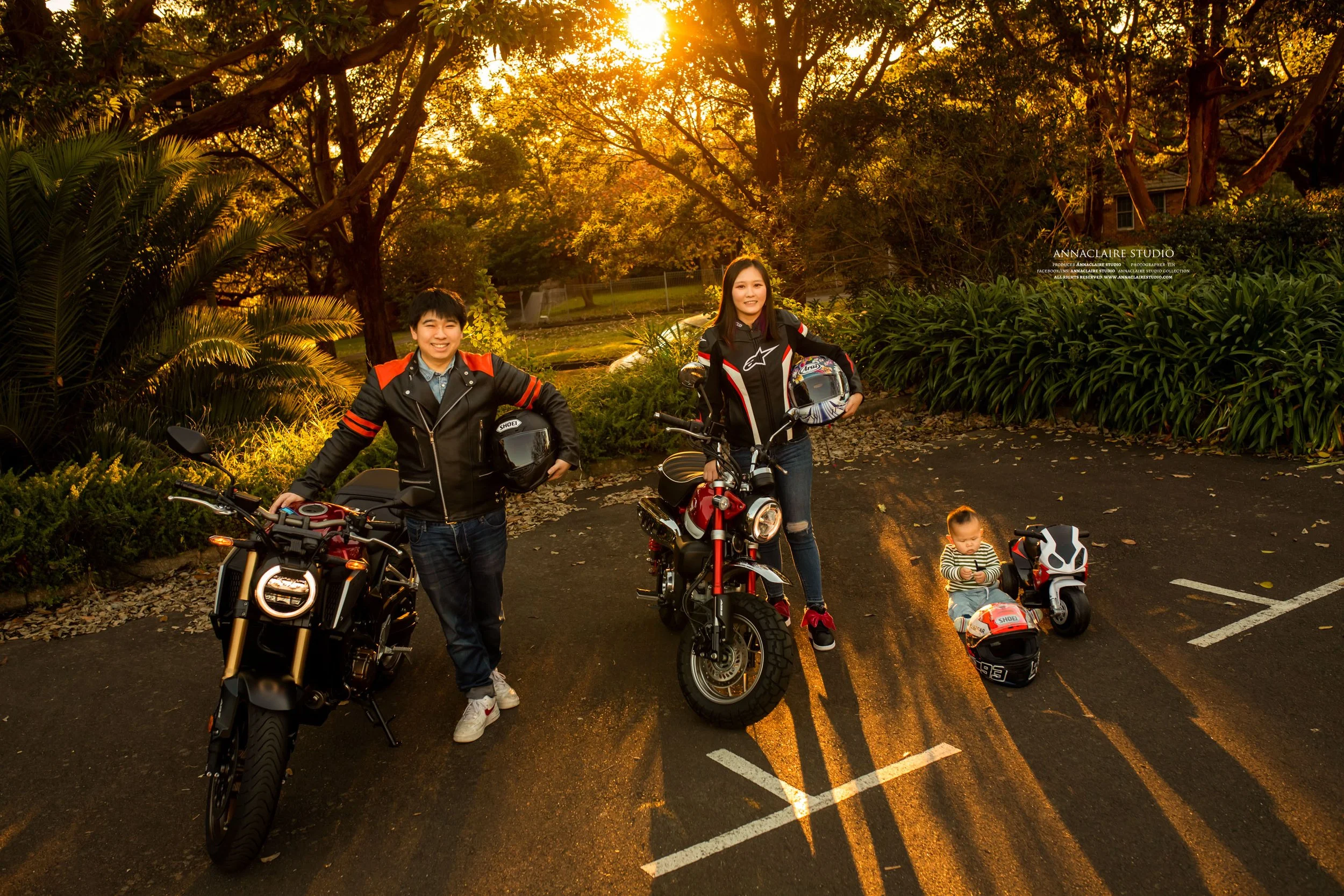 A young boy and girl standing next to motorcycles in a parking lot during sunset. The boy leans on a black motorcycle, and the girl sits on a red motorcycle, holding a helmet. A small child is seated on a toy motorcycle on the ground. Surrounding tre