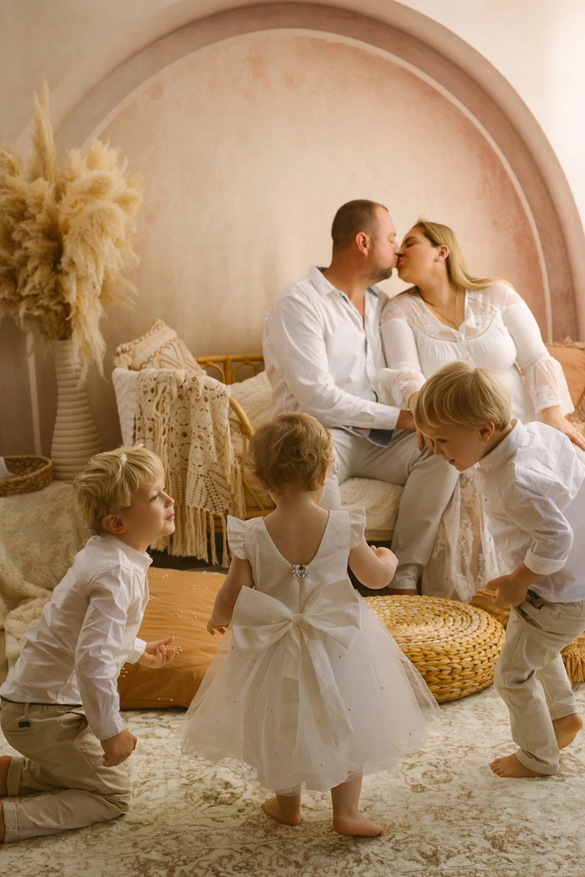 A family of five dressed in white, with the parents sharing a kiss on a bed while the three children, two boys and a girl in a white dress, play on the floor in a cozy, softly decorated room.