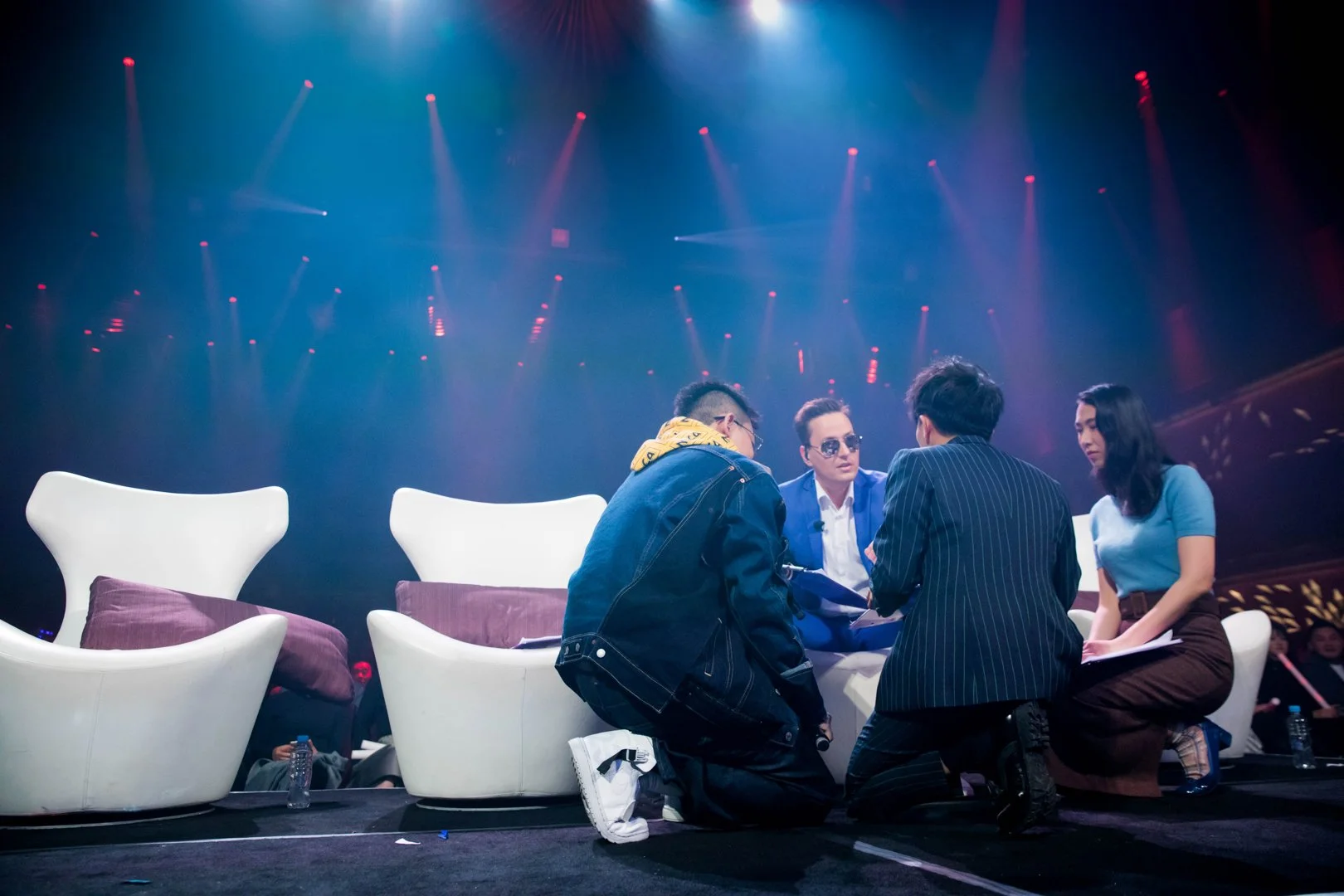 Four people seated on the floor having a discussion, with chairs behind them and a stage with colorful lights in the background.
