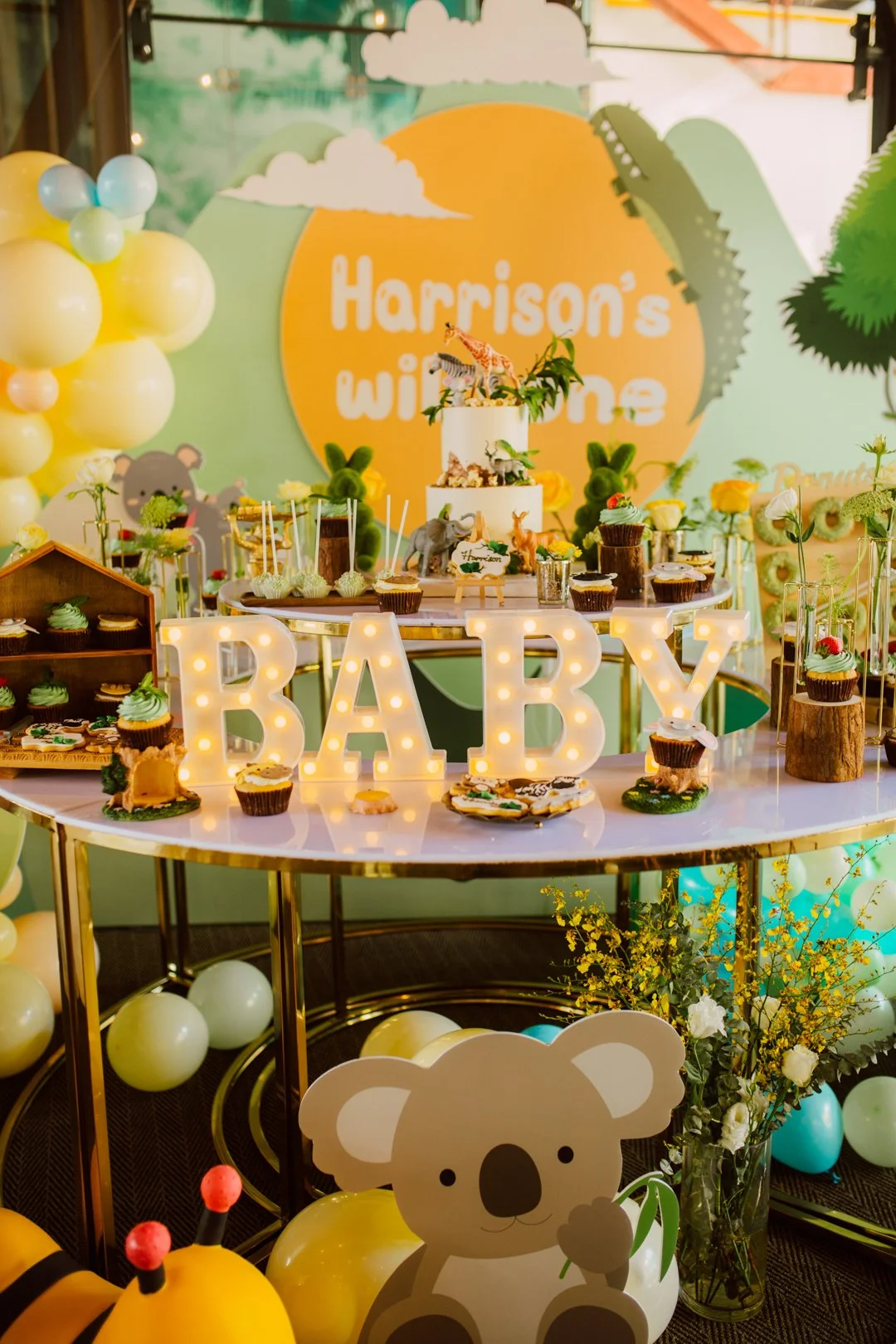Decorated baby shower table with a sign spelling BABY in illuminated letters, animal themed cupcakes, a multi-tiered cake with animal figurines, balloons, and a background featuring a sign that reads "Harrison's Winnie" with jungle and animal motifs.