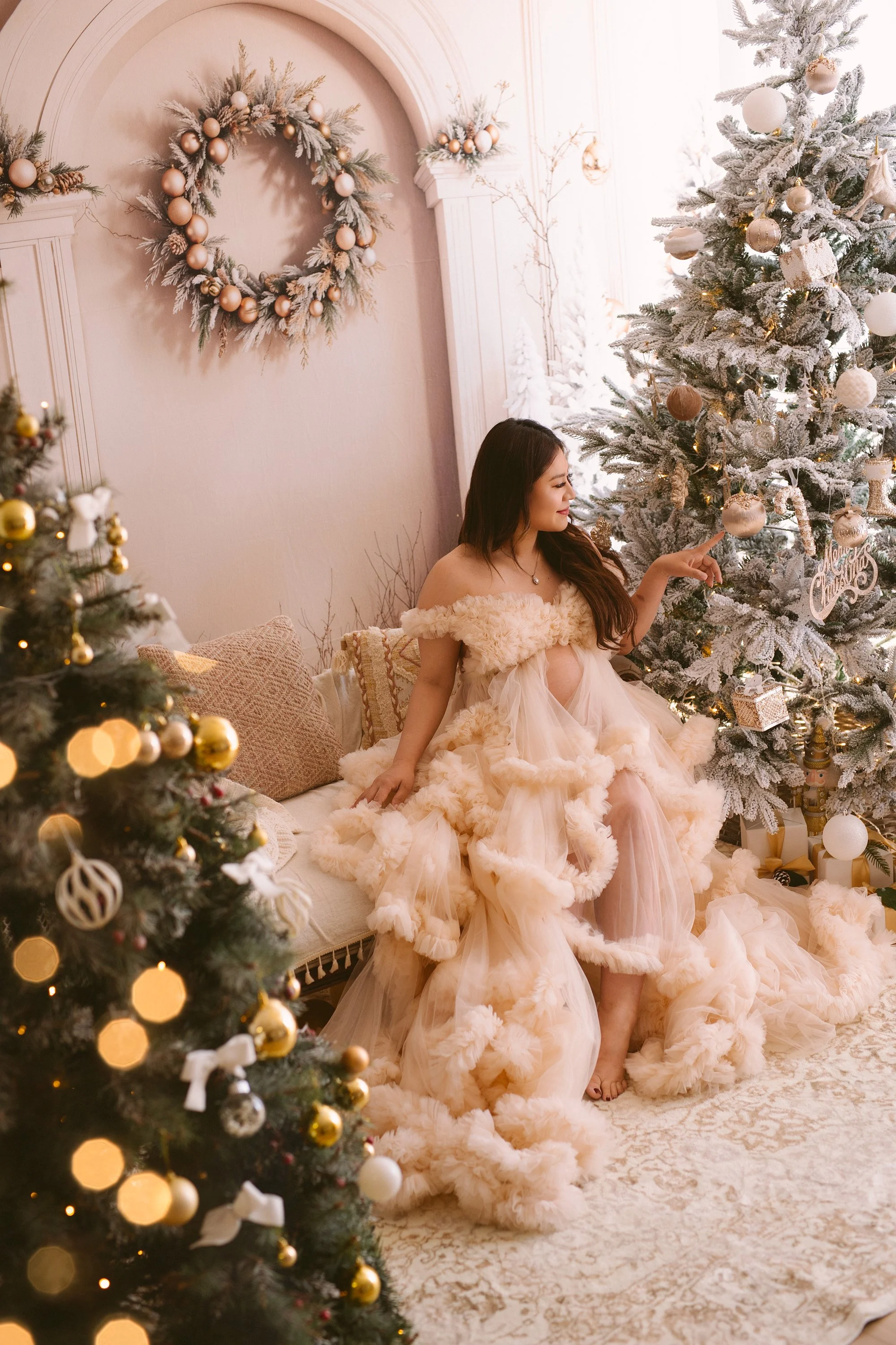 A pregnent lady in a cream-colored, fluffy, off-the-shoulder dress sits on a vintage-style sofa next to a decorated Christmas tree, pointing at an ornament. The room features holiday decorations including a large wreath and candles.