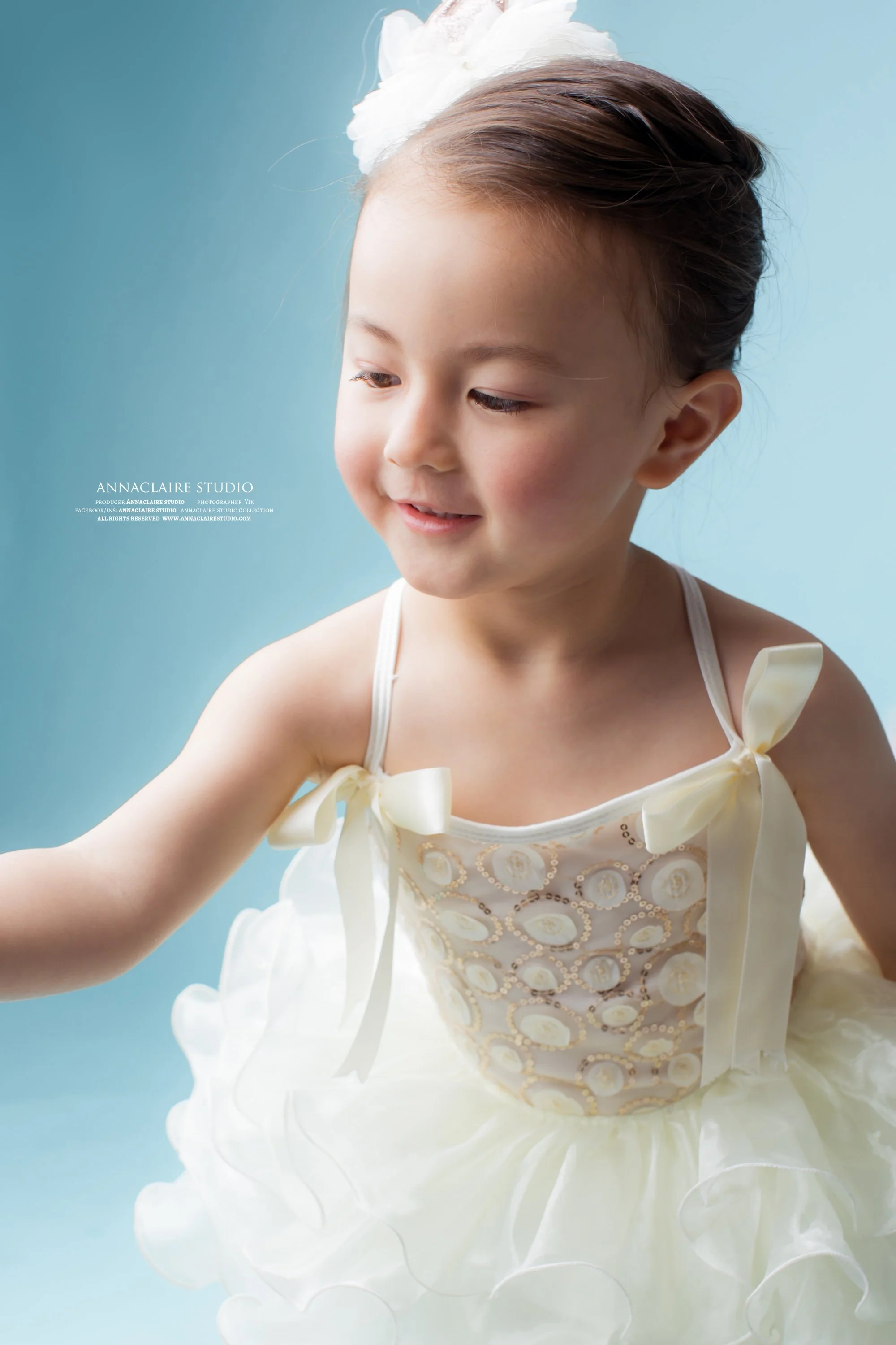 A young girl wearing a white dress with ribbon tie straps and a fluffy skirt, smiling with her head slightly turned to the side against a light blue background.