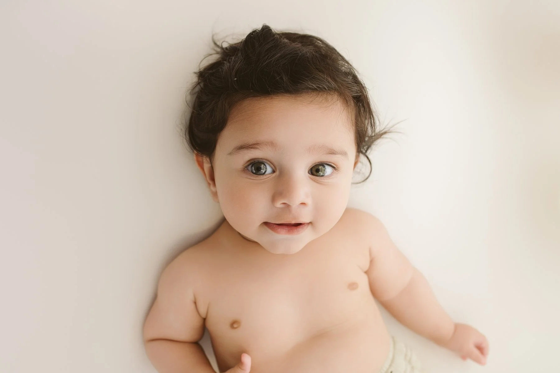 A baby with brown hair and big eyes lying on a white surface, looking up.