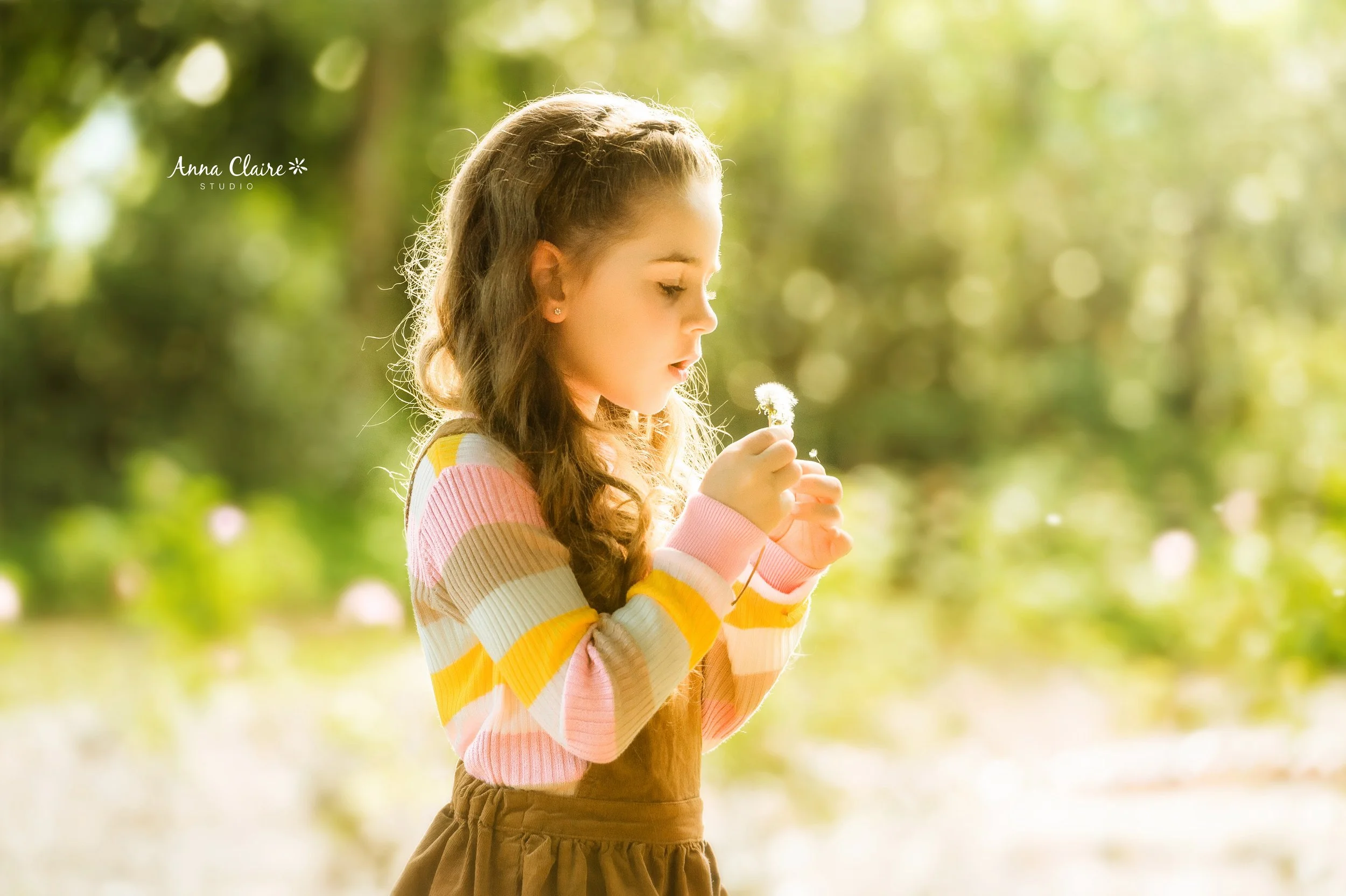 A young girl with long curly hair holding a dandelion flower in a sunny outdoor setting with blurred green trees in the background.