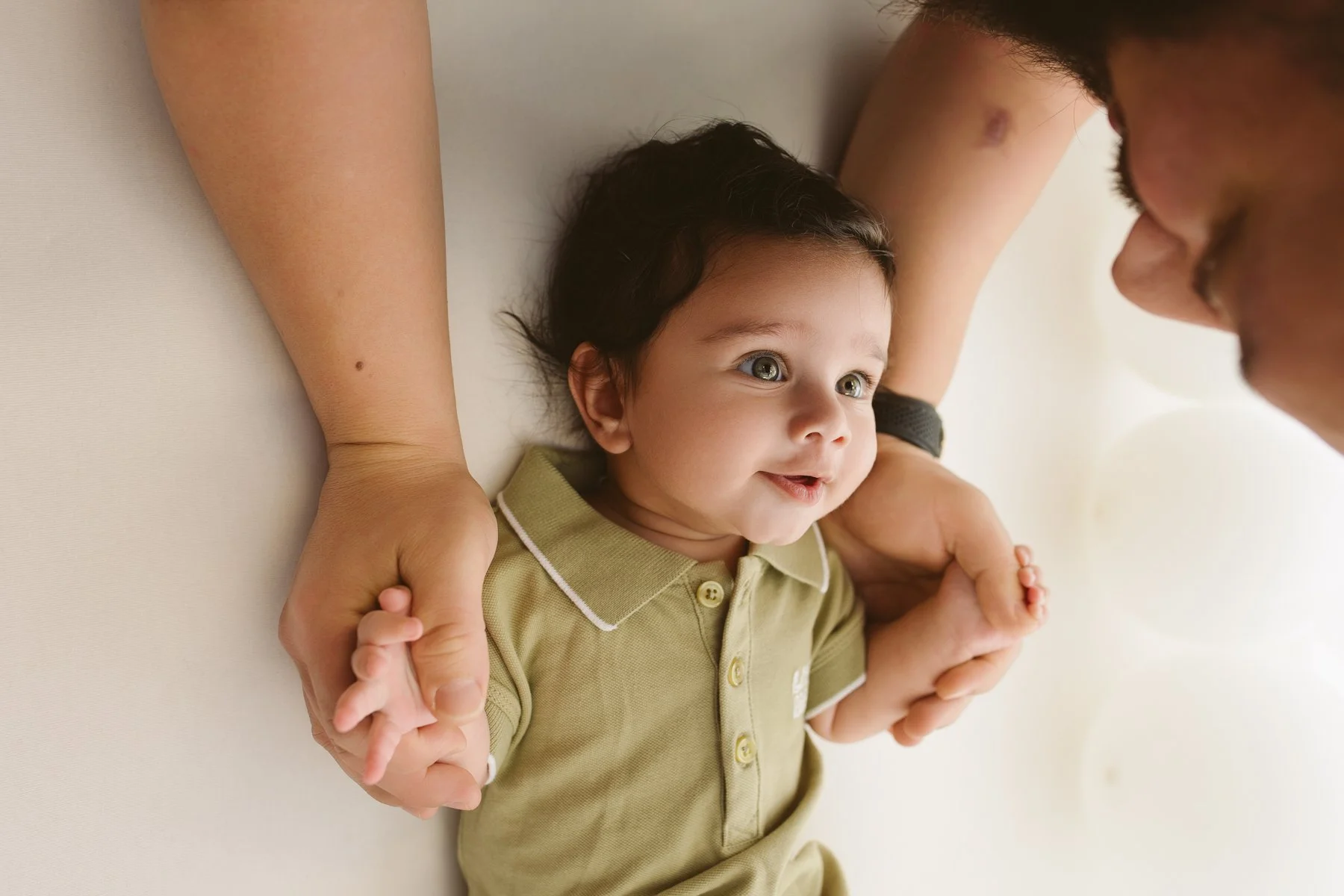 A baby lying on its back, holding hands with an adult, looking up and smiling.