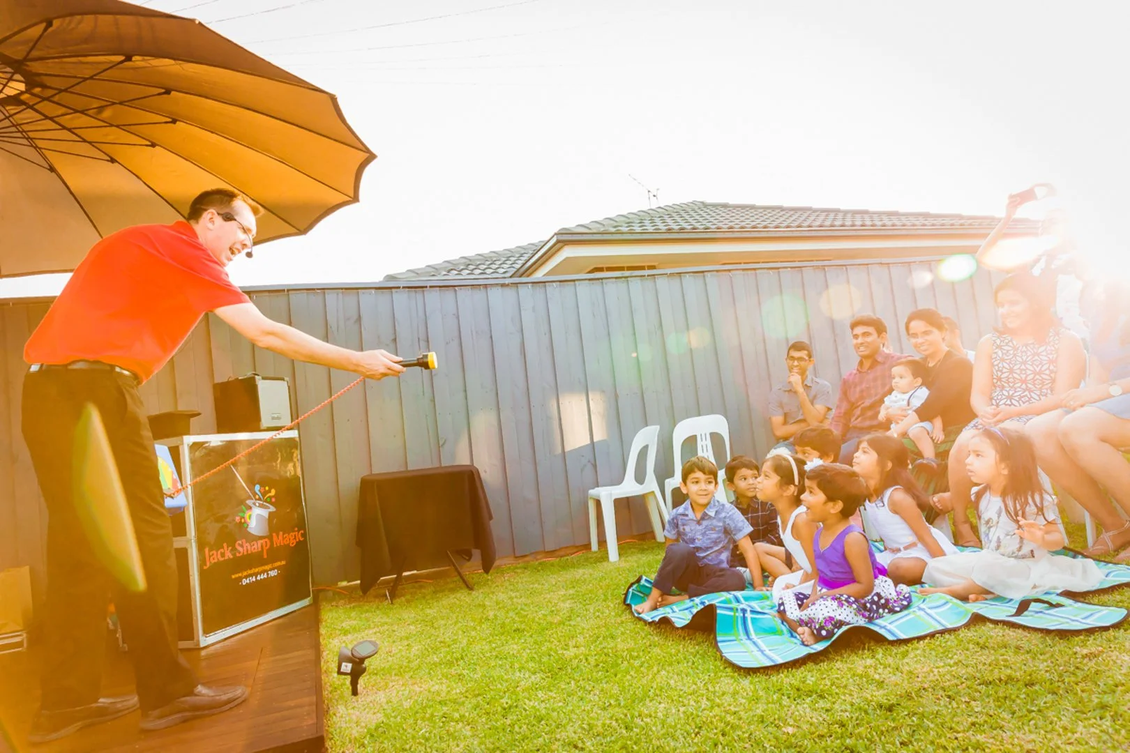 A man in a red shirt performs magic for an audience of children and adults sitting on a blanket and chairs in a backyard, with a sunny sky and a gray wooden fence in the background.