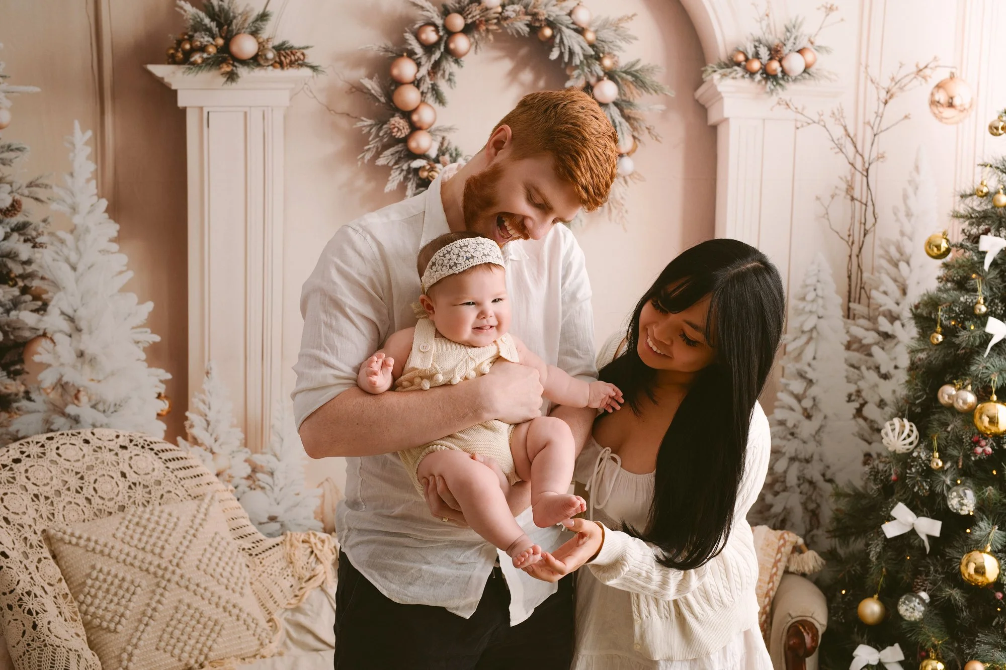 Family celebrating Christmas with a decorated tree and snowy backdrop, with a man, woman, and baby smiling and enjoying the moment.