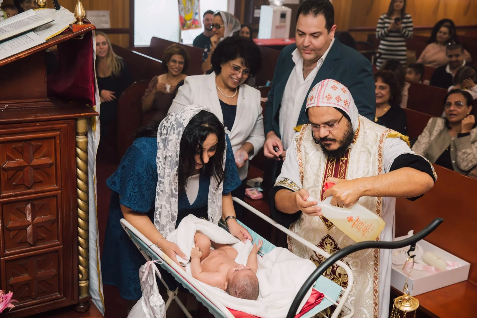 A baptism ceremony in a church with a woman holding a baby in a bassinet, a priest pouring water over the baby's forehead, and several people watching and smiling.